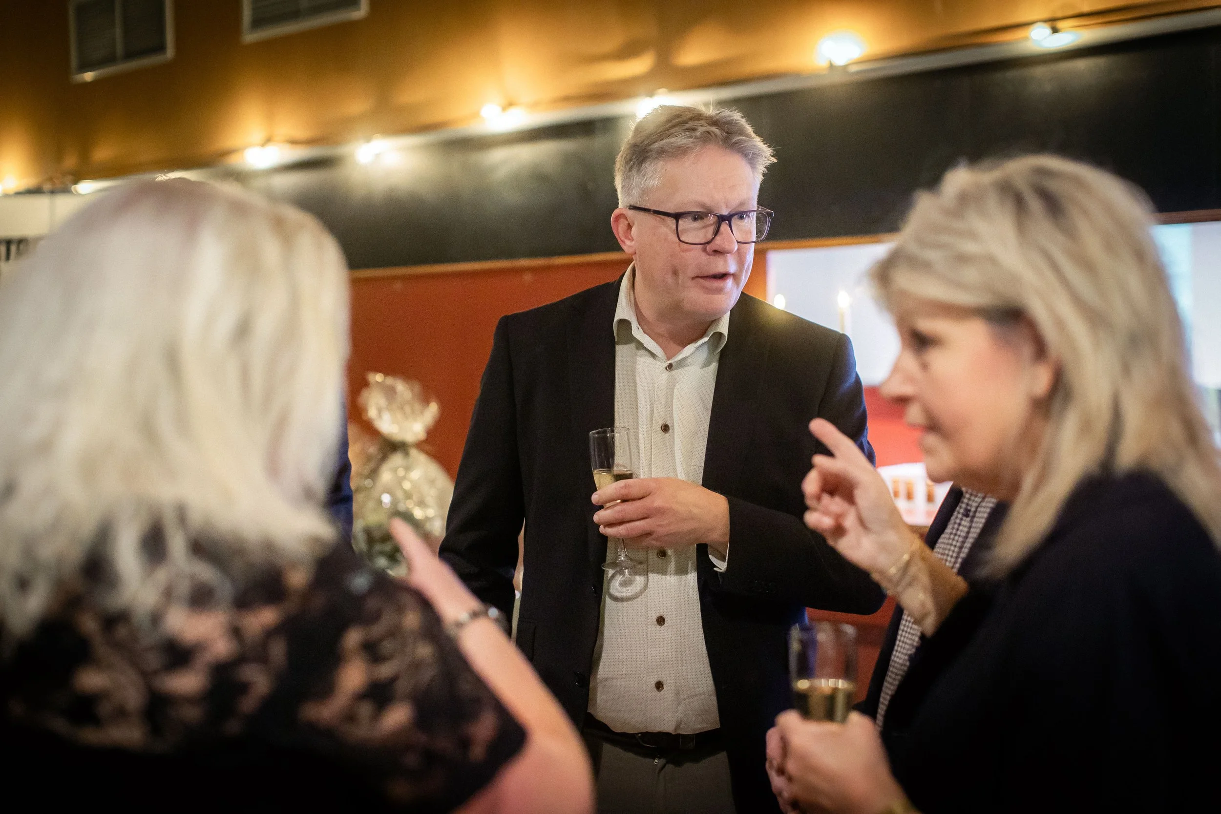 A man in a suit and glasses holding a glass of champagne engaged in conversation with women at a social gathering in a dimly lit room.