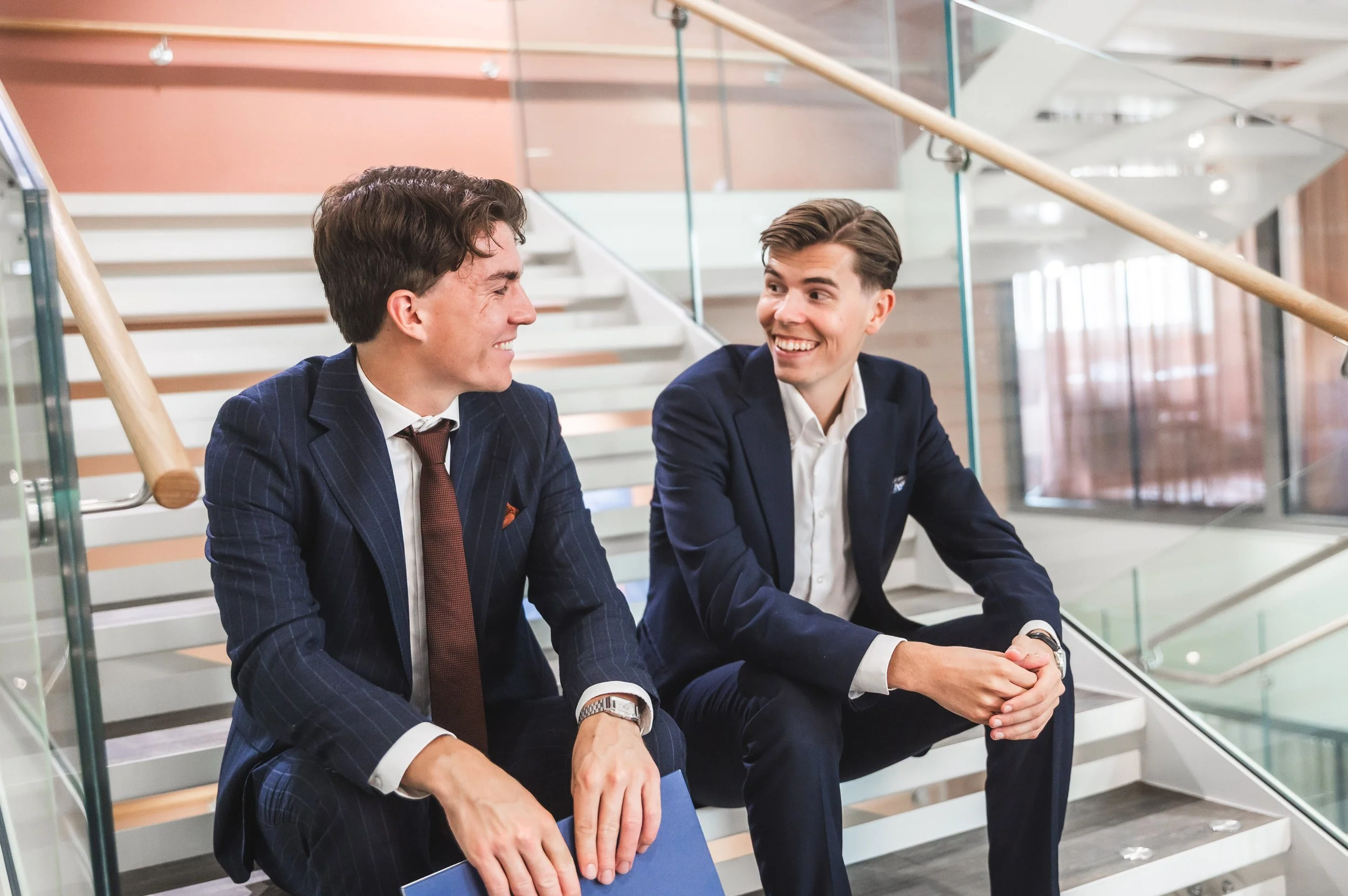 Two young businessmen in suits sitting on a staircase, smiling and talking in a modern office building.