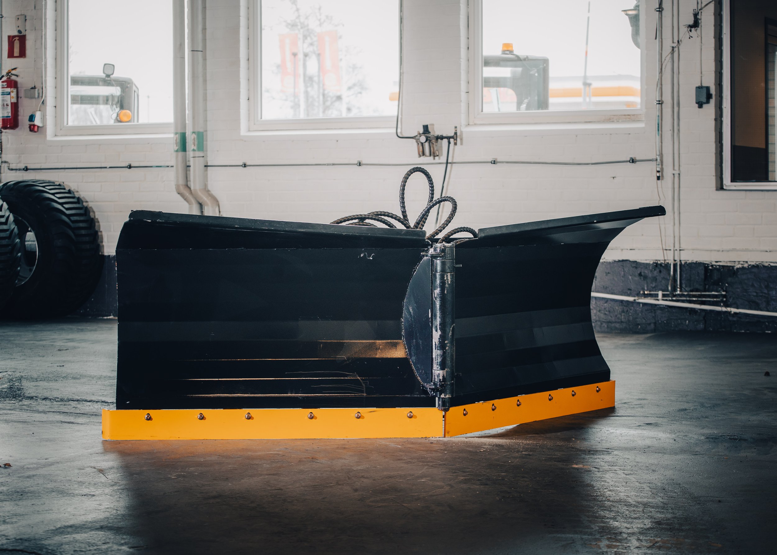 A black snow plow with a yellow bottom edge is inside a garage or workshop. There are large tires in the background, a fire extinguisher on the wall, and windows letting in natural light.