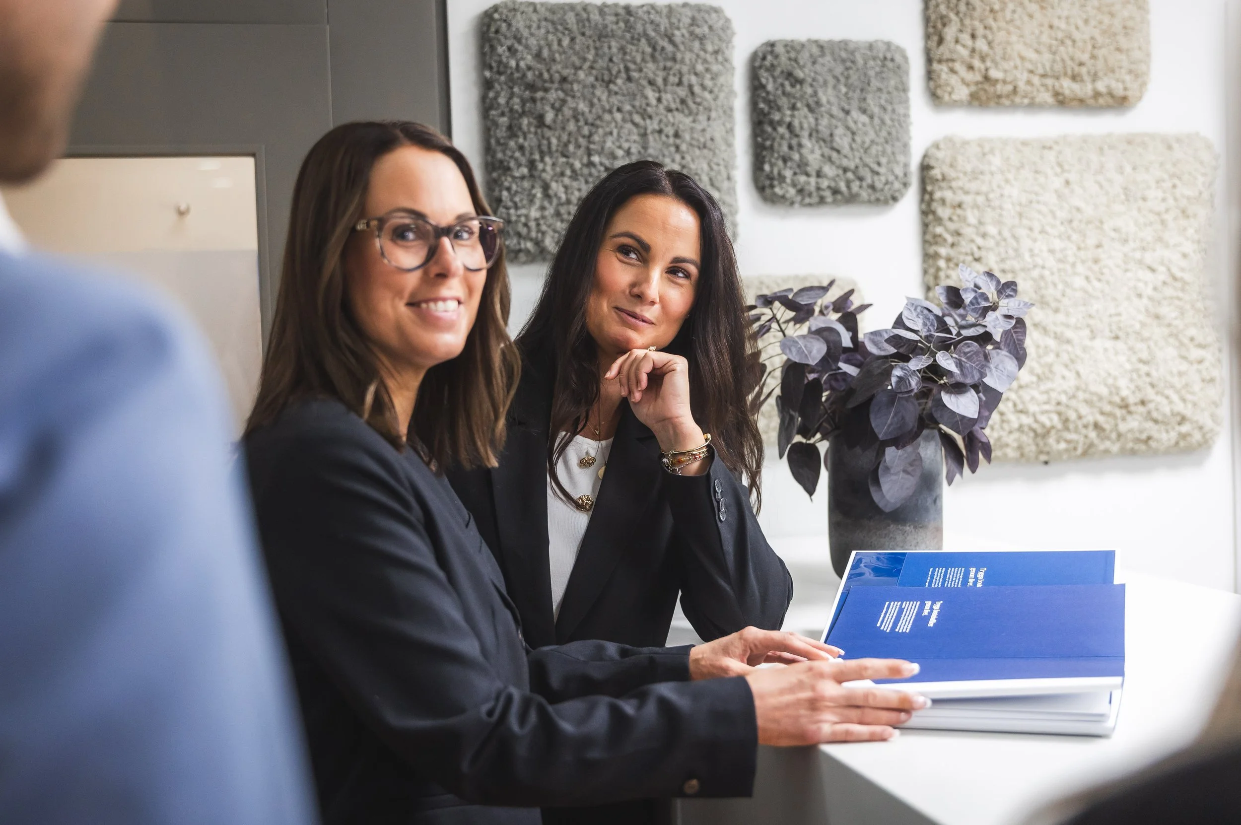 Two women sitting at a table in a professional setting, smiling and engaging in a conversation, with a large flower arrangement and textured wall art in the background.