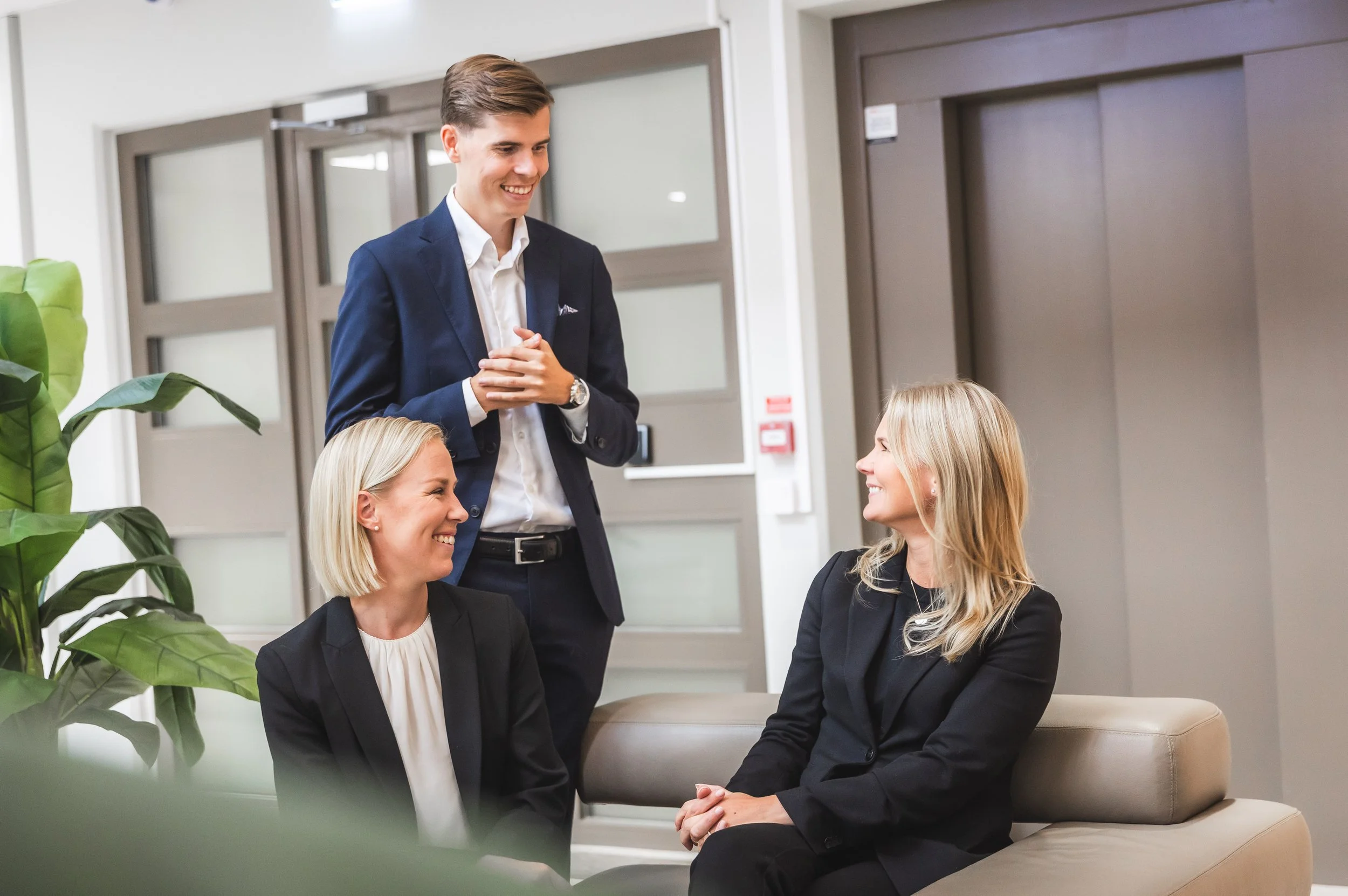 Three business professionals, two women and one man, engaged in a friendly conversation in an office lobby. The man is standing, smiling, and wearing a navy blue suit, while the women sit on a beige sofa, smiling and dressed in black blazers.