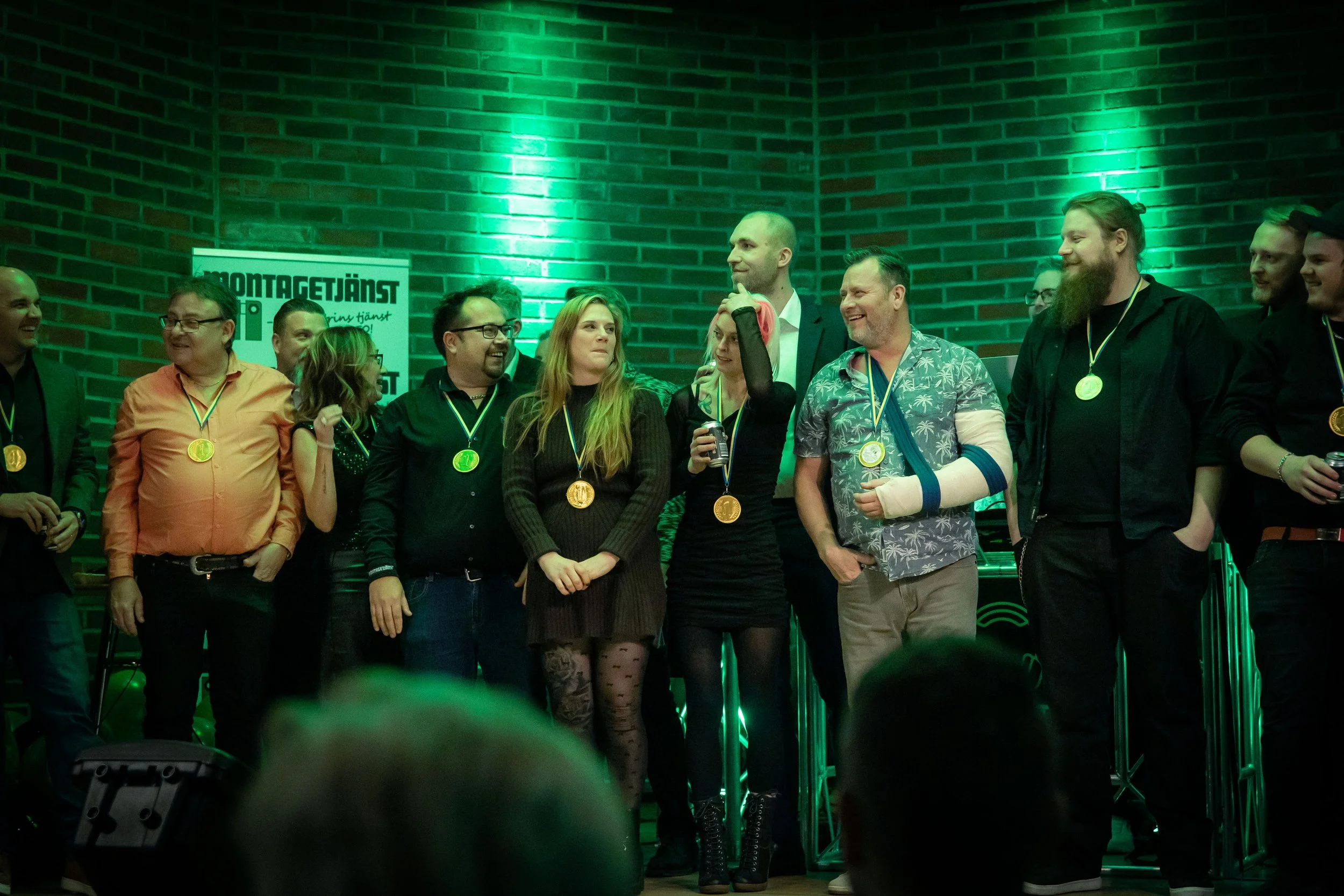 Group of people wearing medals on stage during celebration or award ceremony, with a brick wall background and green lighting.