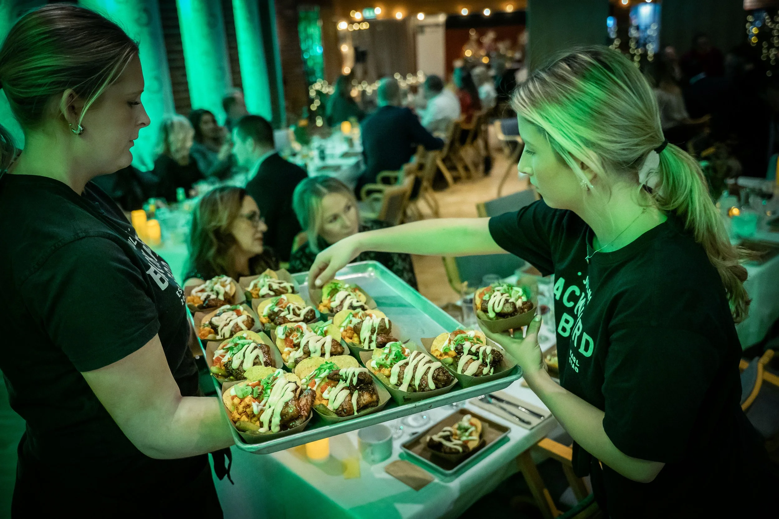 Two women serve loaded hot dogs with toppings at a banquet or event with many guests seated at tables in the background.