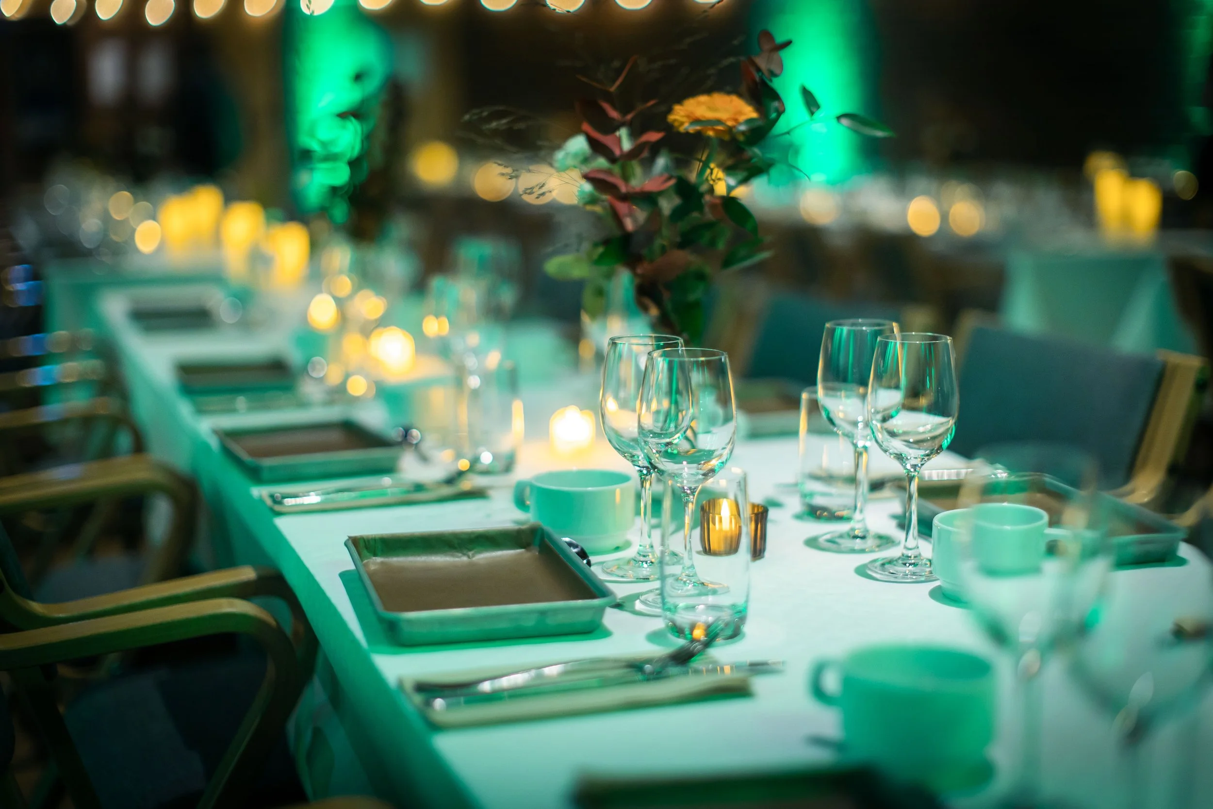 Set dining table with wine glasses, cups, and plates, decorated with a floral centerpiece and candles, in a dimly-lit restaurant.