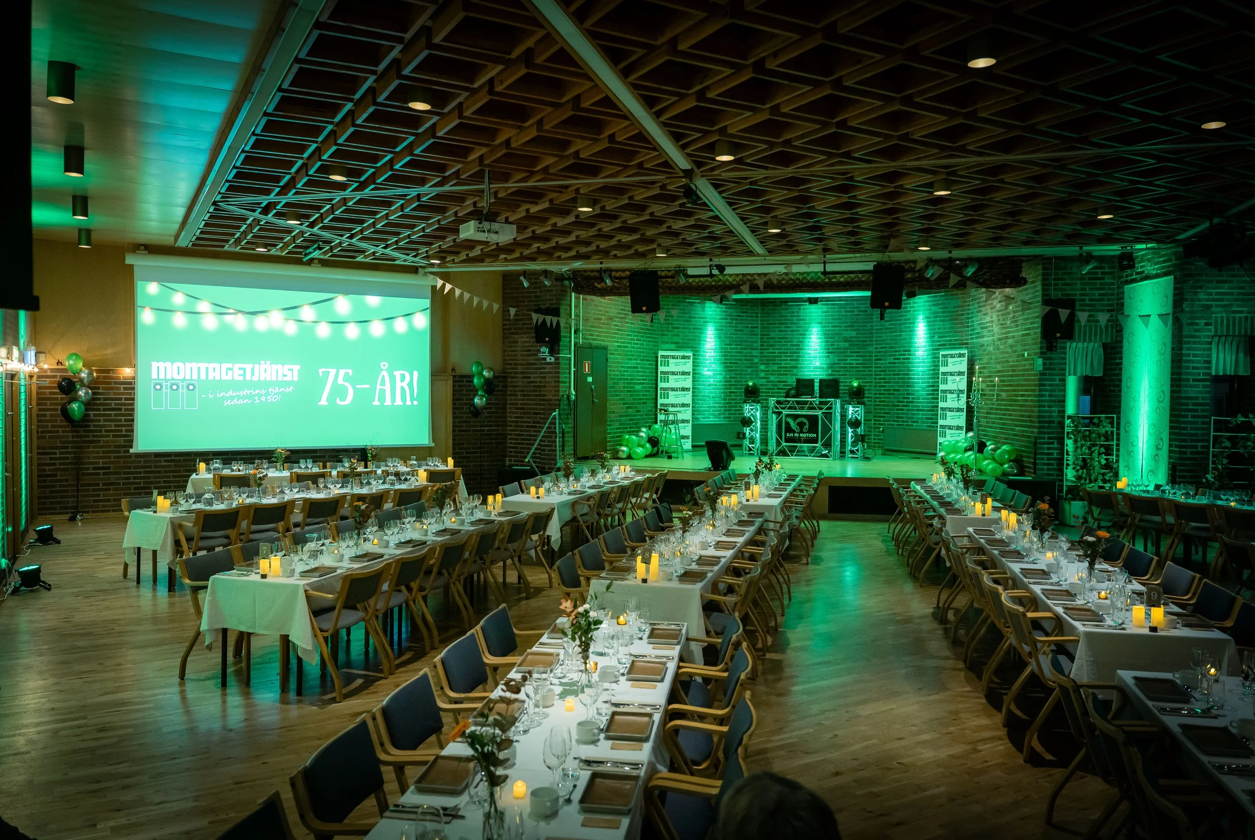 Event hall decorated with green lighting, long tables set with plates, glasses, and candles, stage with DJ equipment and balloons, and a large screen displaying a celebration message.
