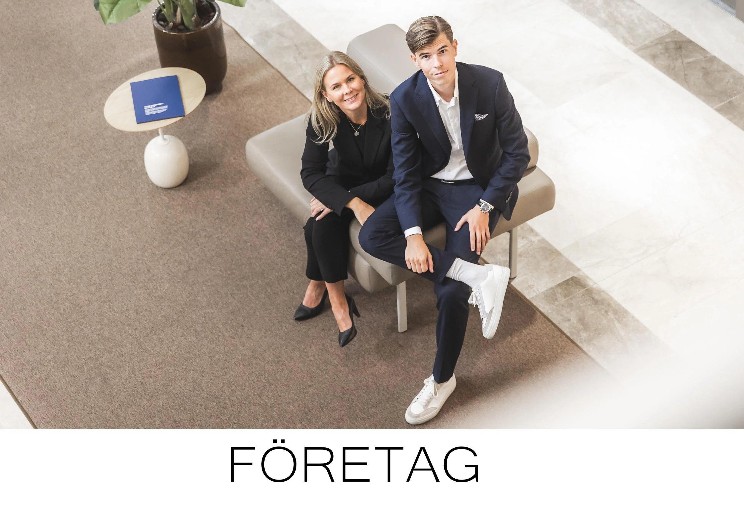 Two well-dressed people — a woman and a young man — sitting on a bench in an indoor setting, looking up at the camera. The woman is wearing a black outfit with black heels, and the young man is in a blue suit with white sneakers. There is a small rou