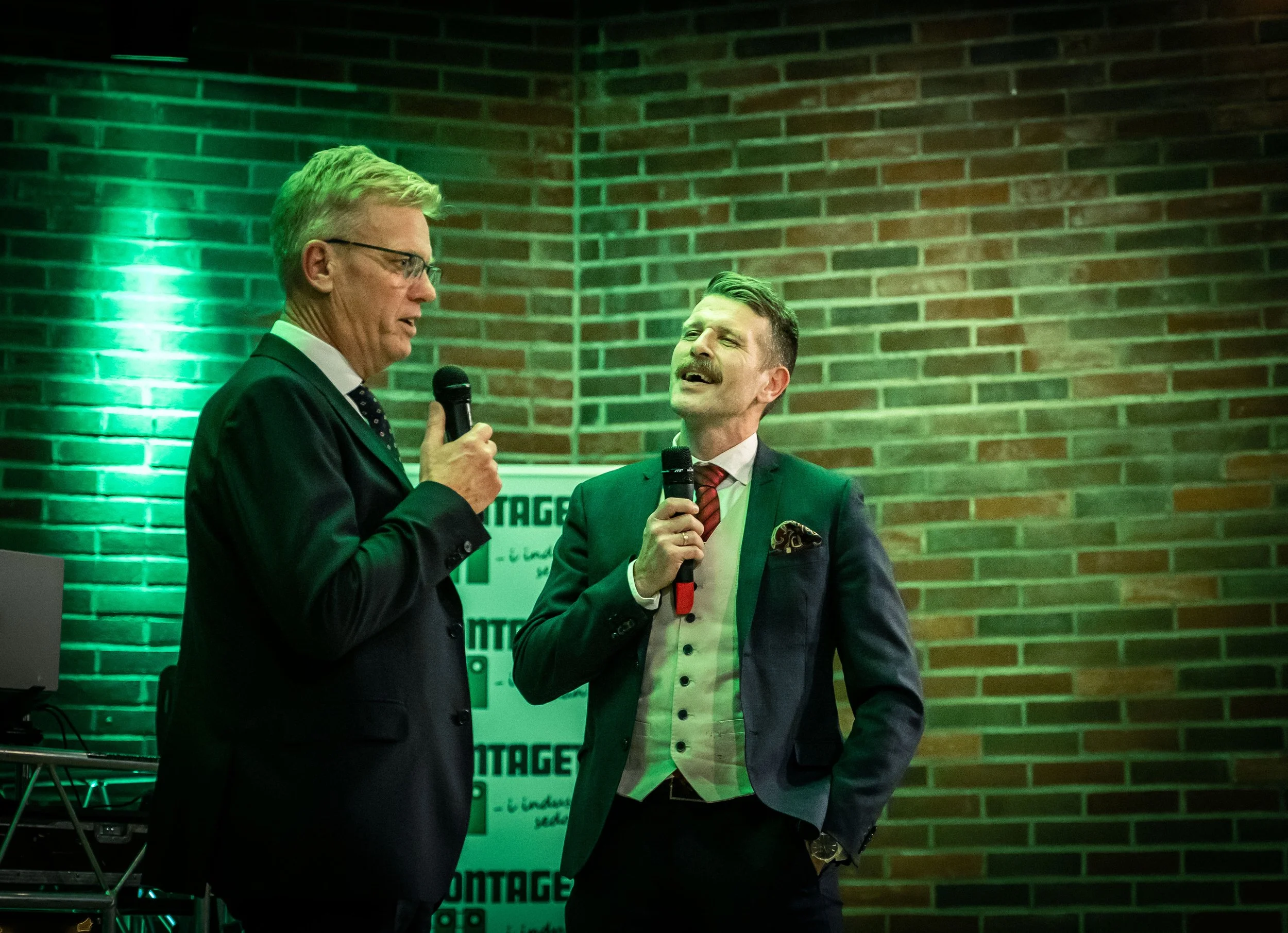 Two men in suits holding microphones, engaged in conversation at an indoor event. The background features a brick wall and a poster or banner with partially visible text. Green lighting illuminates part of the scene.