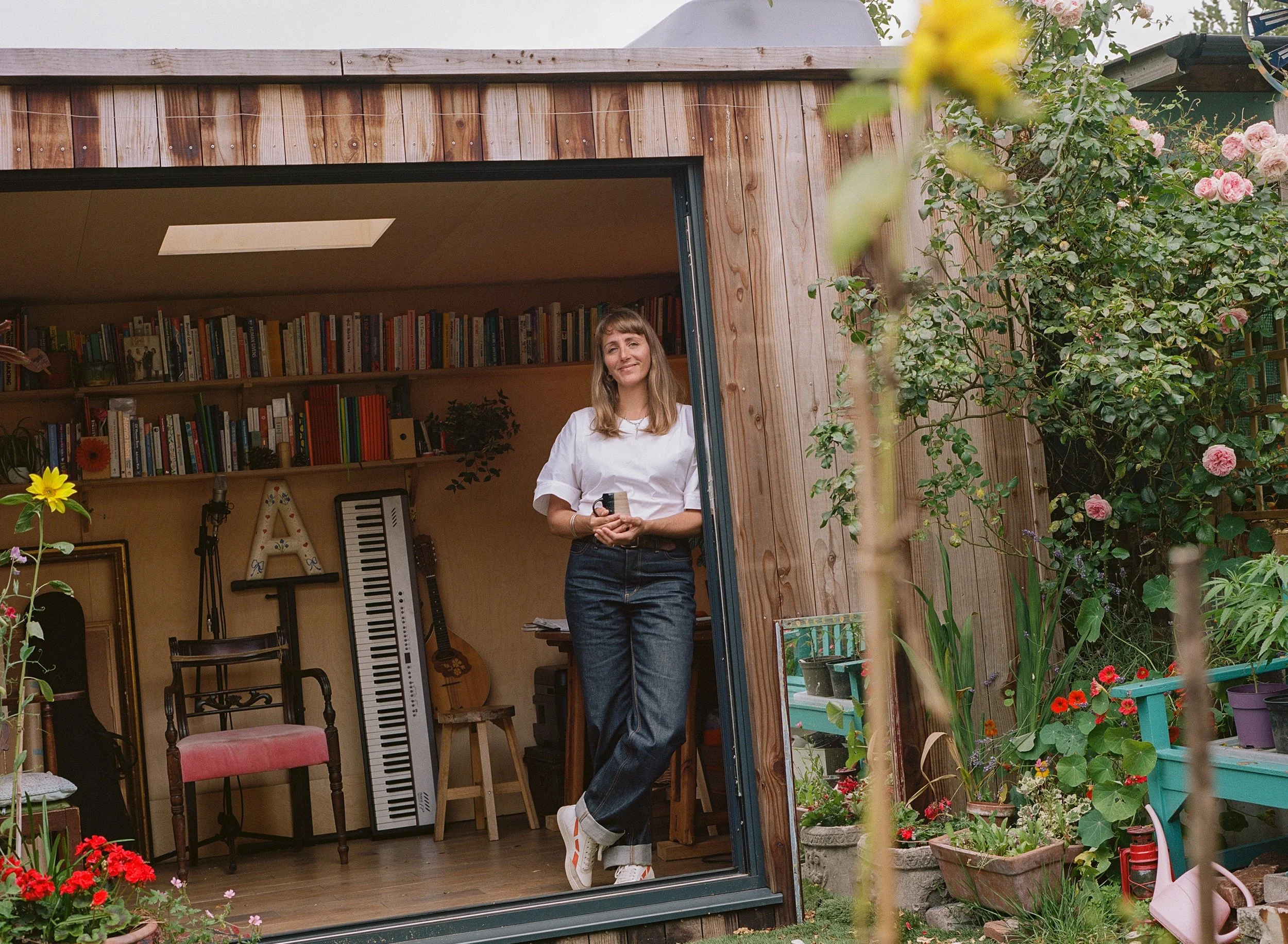 A woman with long hair, wearing a white shirt and dark jeans, standing inside a small wooden garden shed, smiling at the camera. The shed's interior contains bookshelves, a keyboard, and decorative items. The shed door is open, and the shed is surrounded by colorful flowers and green plants outside.