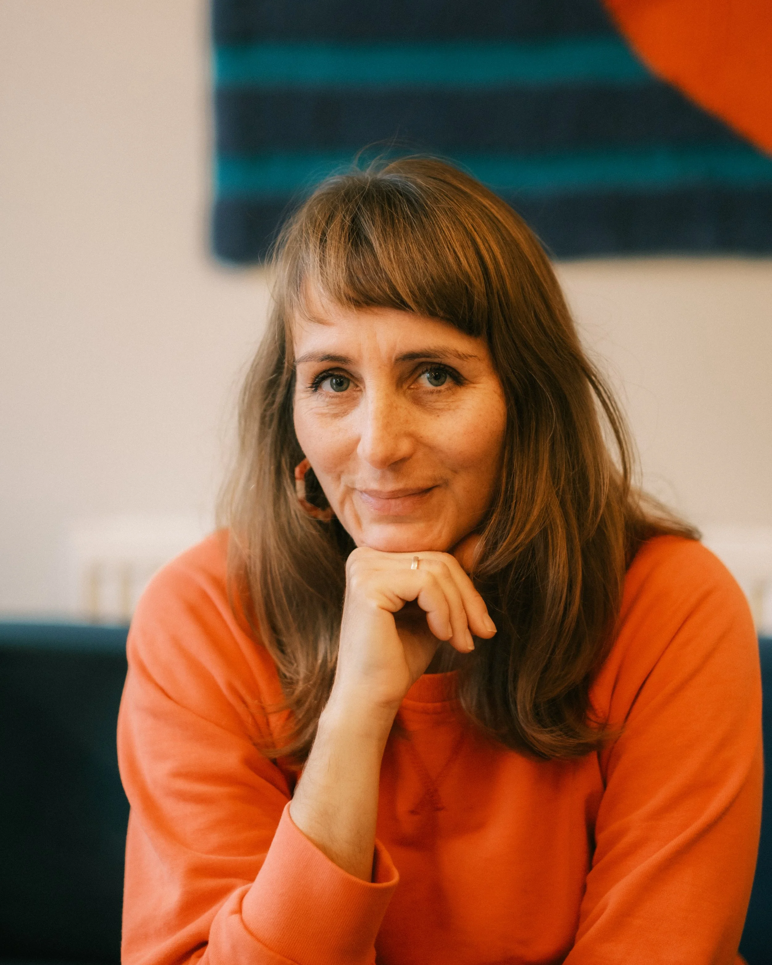 A woman with reddish-brown hair and blue eyes, smiling gently, resting her chin on her left hand, wearing an orange long-sleeve shirt, sitting in an indoor setting with a blurred abstract painting in the background.