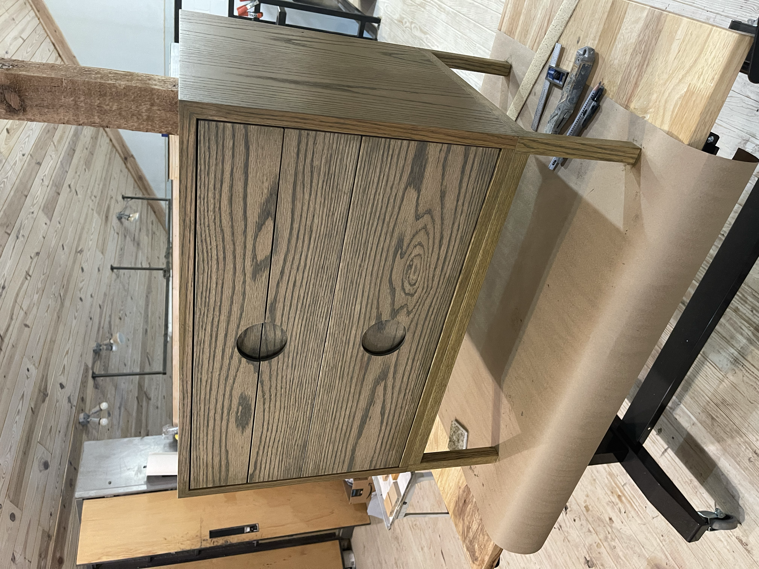 A wooden cabinet with two drawers and two doors, sitting on a work table in a woodworking shop