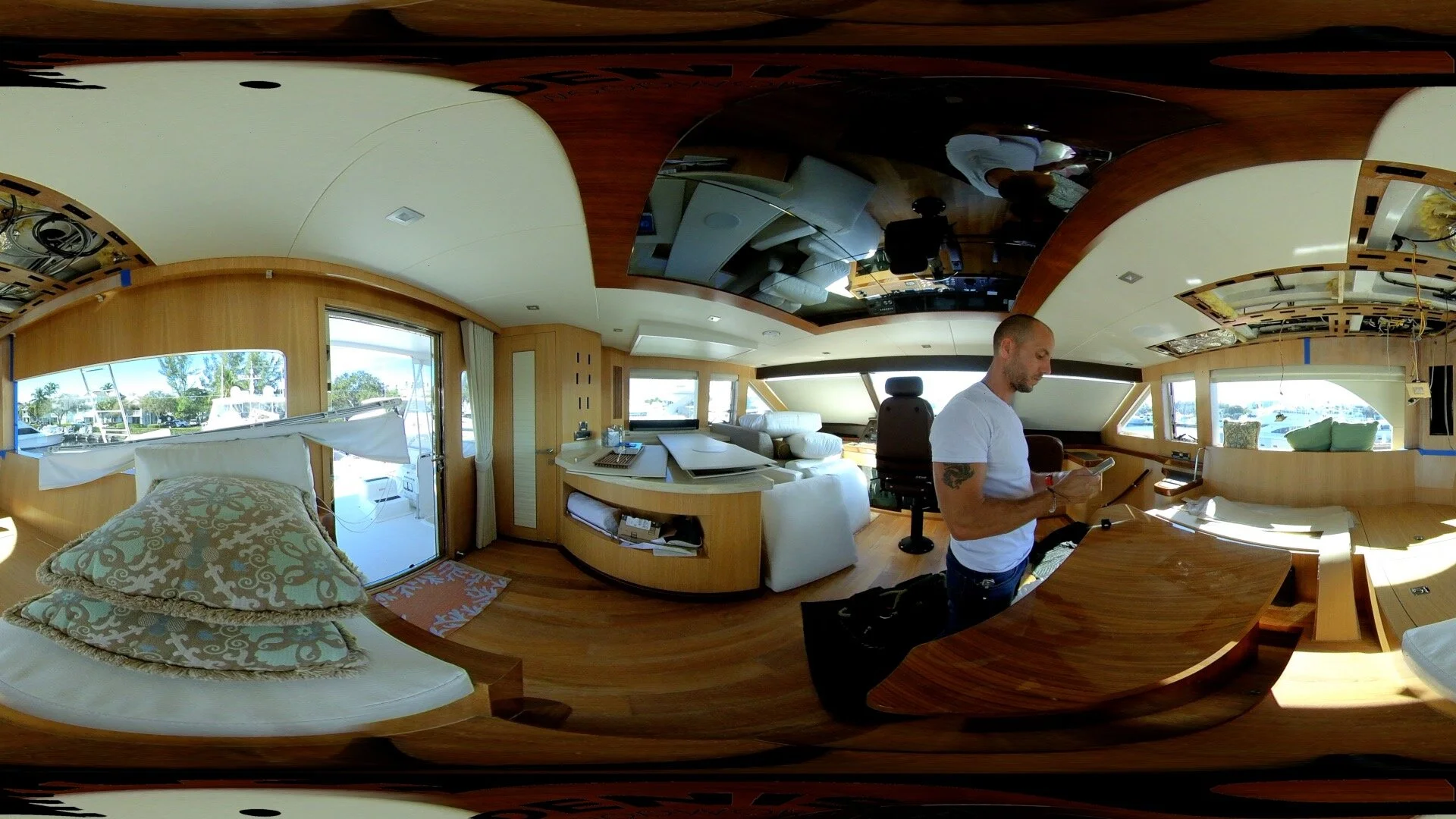 Interior of a boat or yacht with a man standing at a wooden desk, large windows with views of water and boats, and a ceiling still under construction.