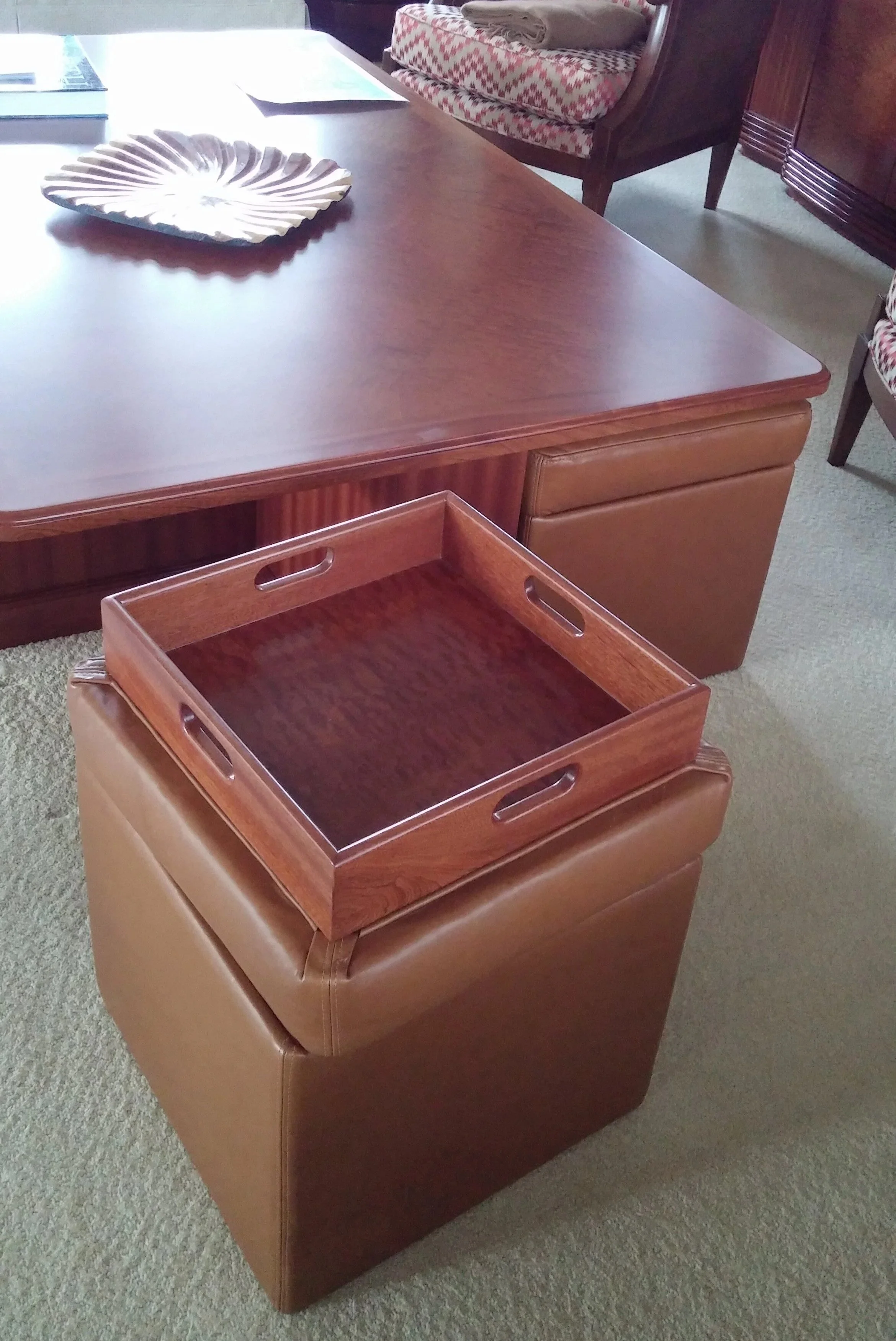 A wooden tray placed on a brown cushioned ottoman in front of a wooden table with a decorative dish and papers, in a room with patterned chairs and a wooden cabinet.
