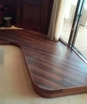 A corner of a room with wooden flooring, a glass sliding door, curtained window, and a beige bed or sofa with wooden trim.