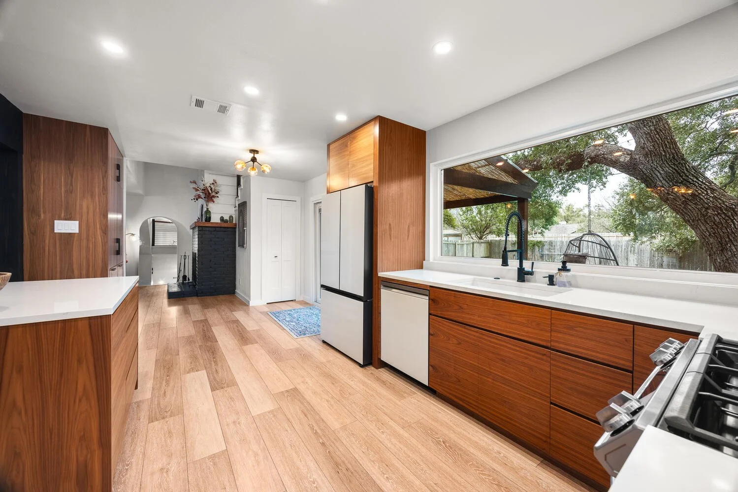 Modern kitchen with wooden cabinets, white countertops, a large window showing trees outside, and a stove in the foreground.