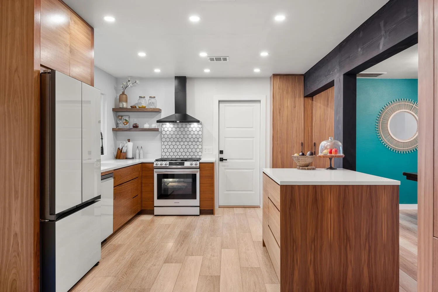 Modern kitchen with wooden cabinets, white countertops, stainless steel appliances including a refrigerator and stove, open shelving with decorative jars, and a teal accent wall with a round sunburst mirror.