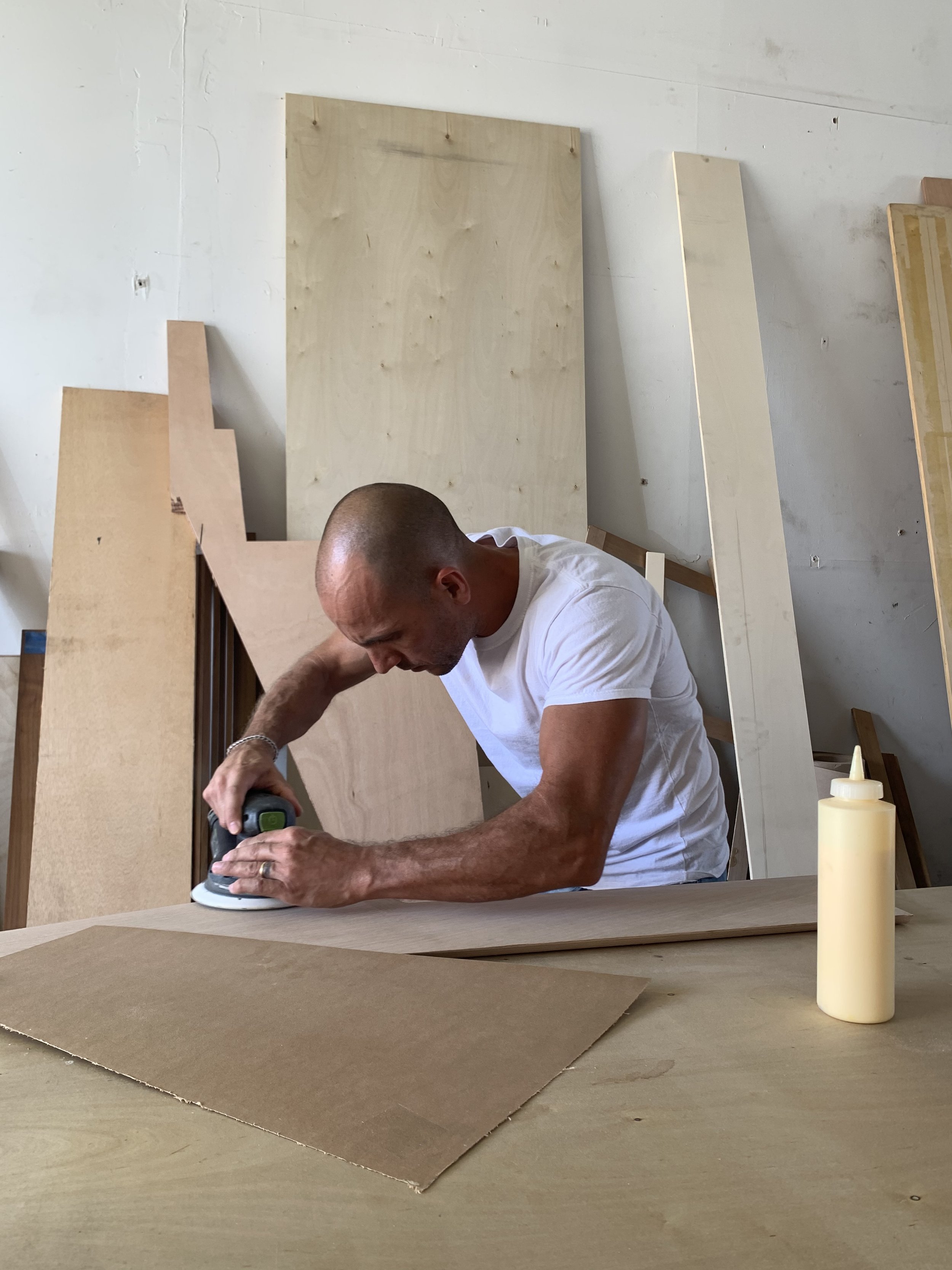 A man in a white t-shirt using a small electric sander on a piece of wood or cardboard in a woodworking shop with large wooden boards and a bottle of glue visible.