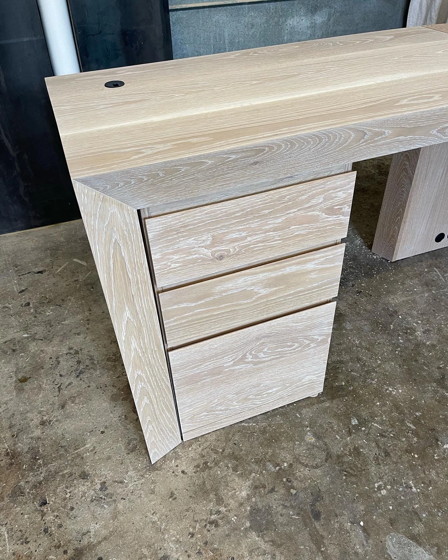 A light-colored wooden desk with four drawers on the left side and a hole on the top surface for cables, placed on a concrete floor.