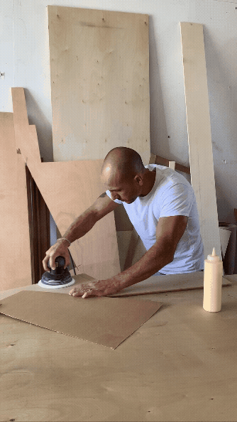 Man in a white T-shirt using an iron on a piece of fabric in a woodworking workshop with large wooden boards in the background.