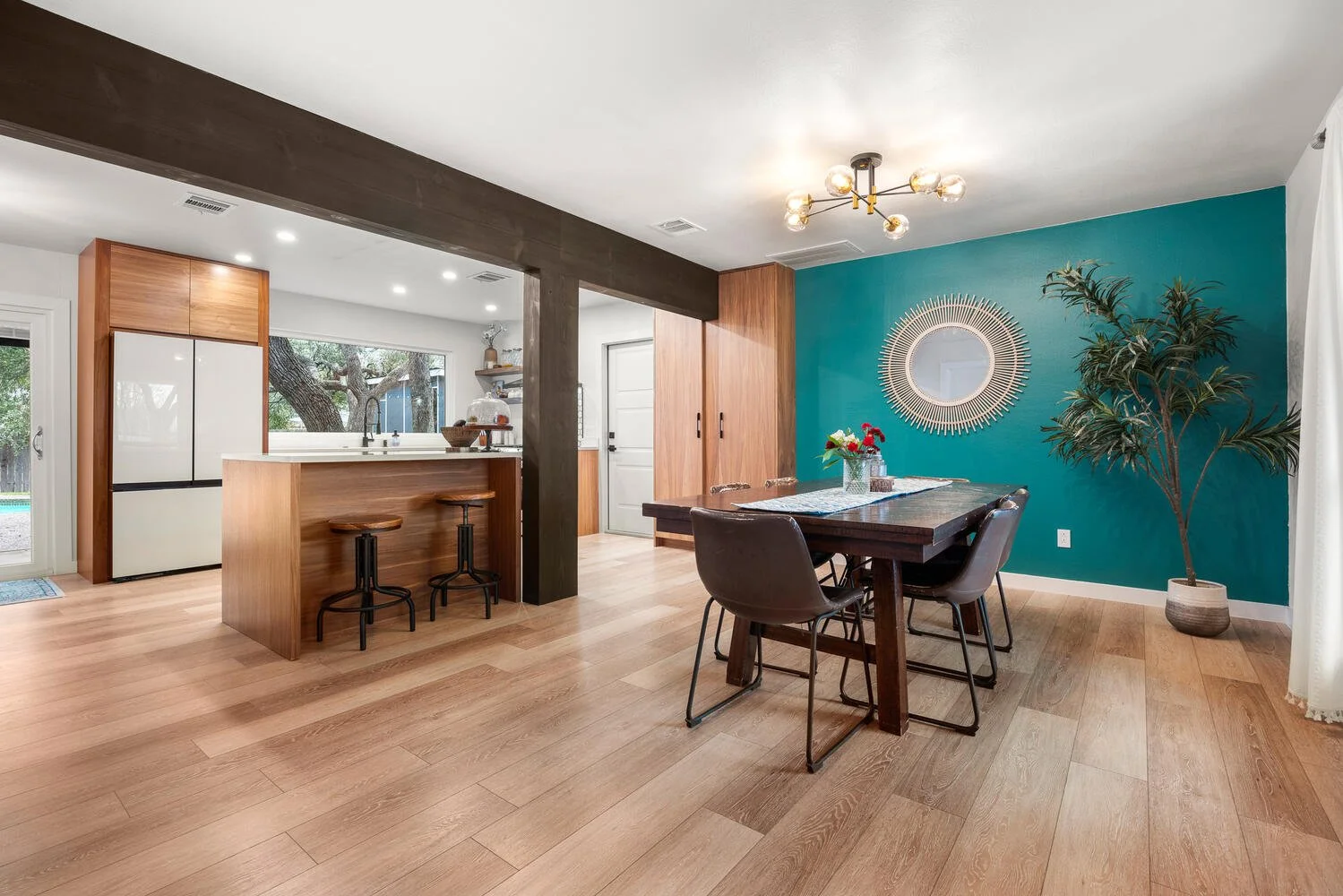 Open-concept dining area and kitchen with wooden floors, teal accent wall, modern chandelier, dark wood dining table with chairs, large potted plant, and a breakfast bar with stools.