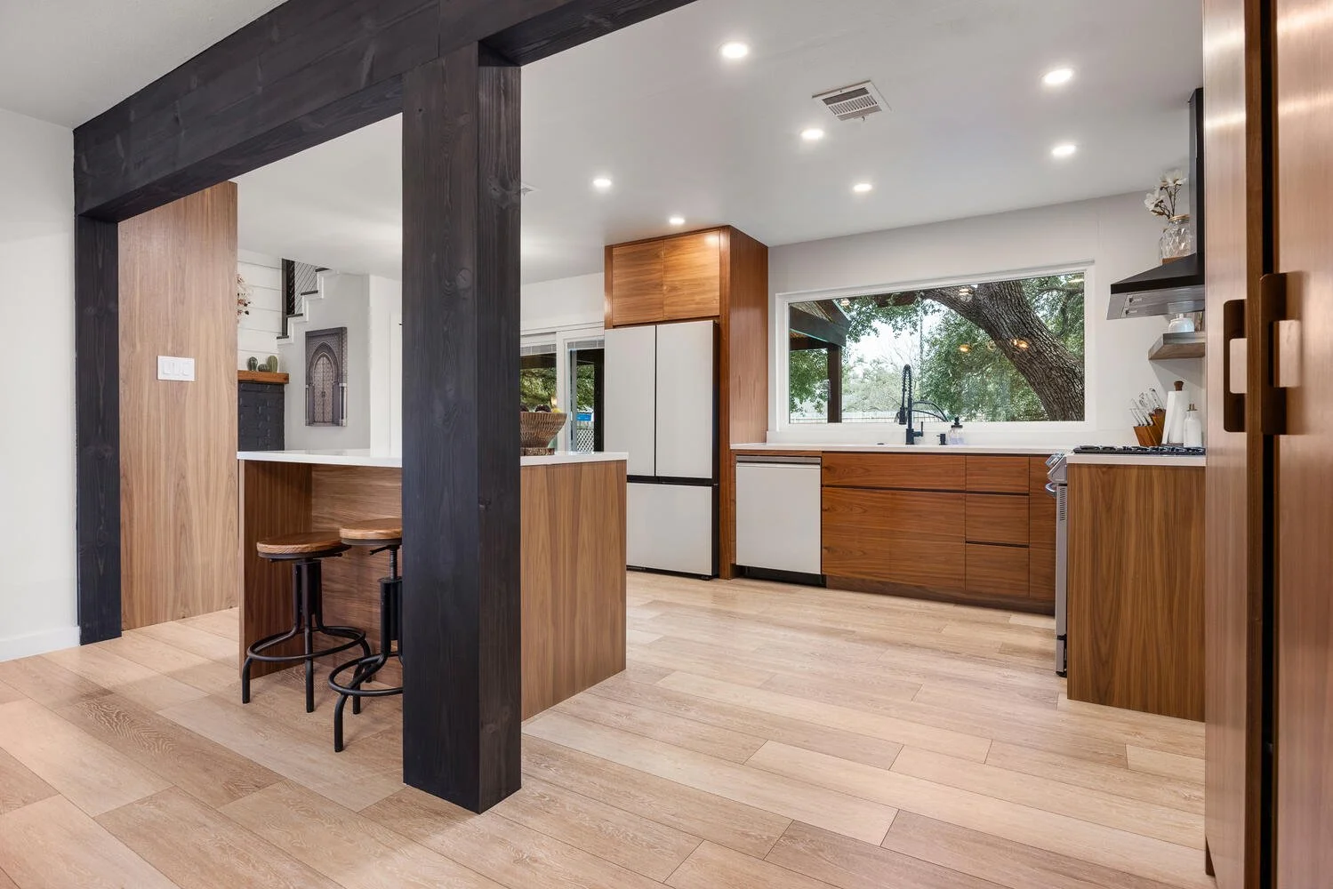 Modern kitchen with wood cabinets, a large window overlooking trees, black fixtures, and a light wood floor.
