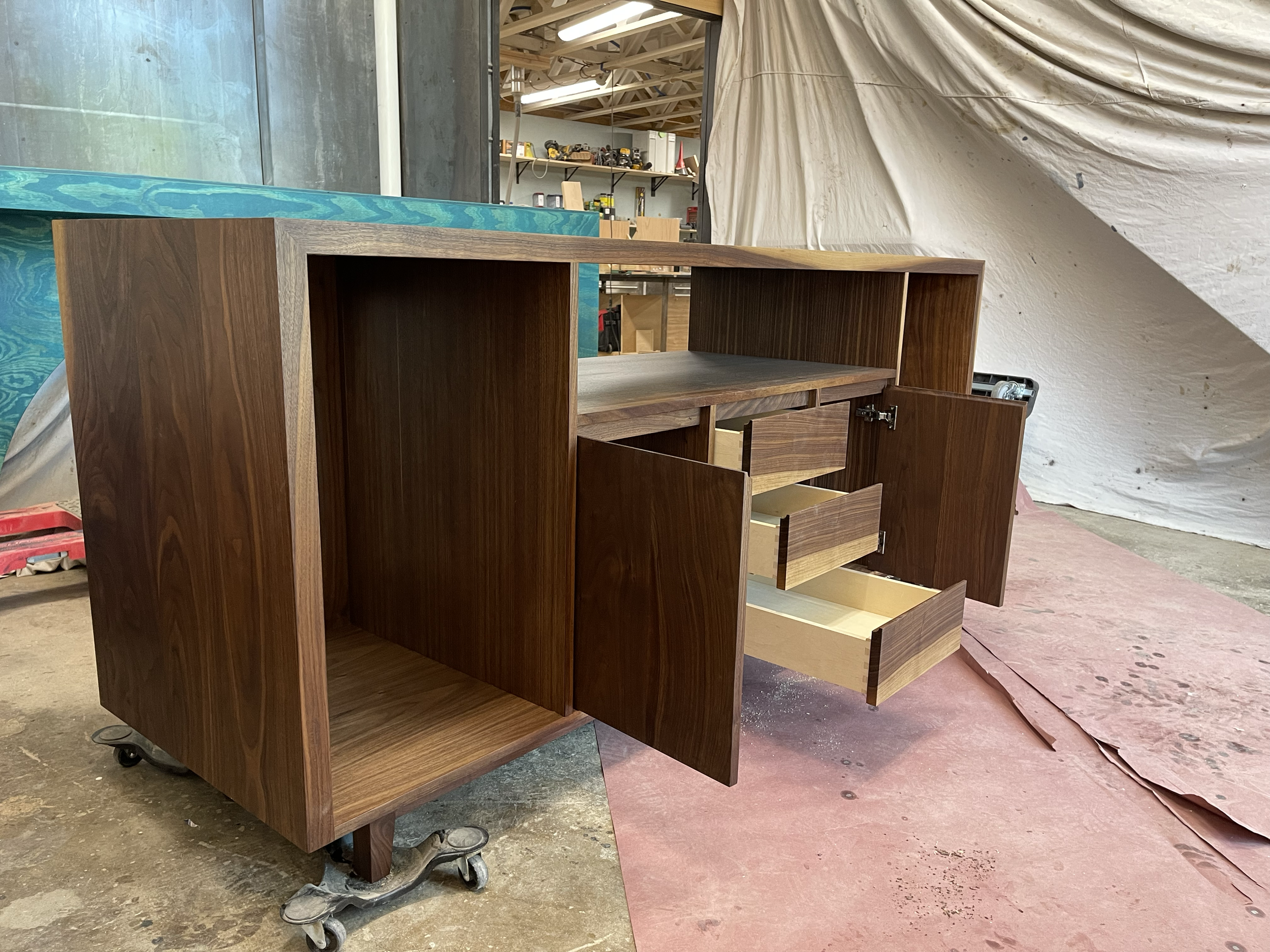 Unfinished wooden cabinet with four drawers and two doors, on wheels in a woodworking shop.