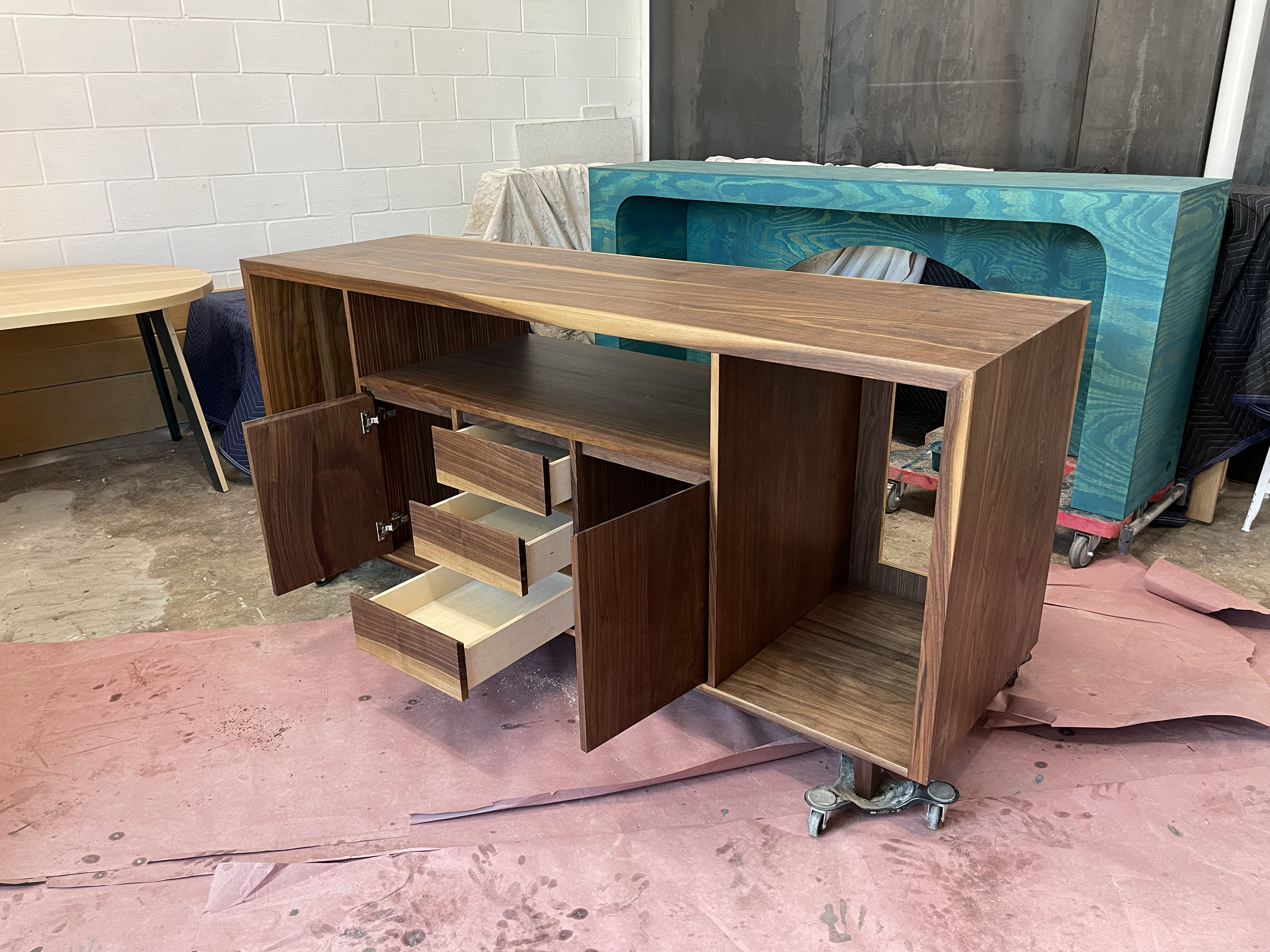 A wooden cabinet on wheels with multiple drawers and open storage compartments, placed on pink protective floor covering in a workshop.