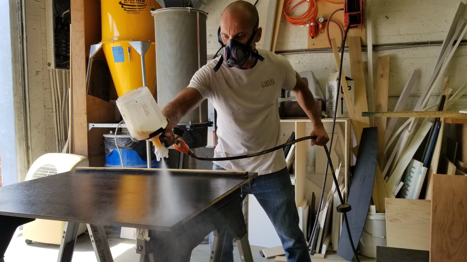 A man wearing a white t-shirt, jeans, and a mask with respirator filters is spray painting a piece of furniture in a woodworking workshop.
