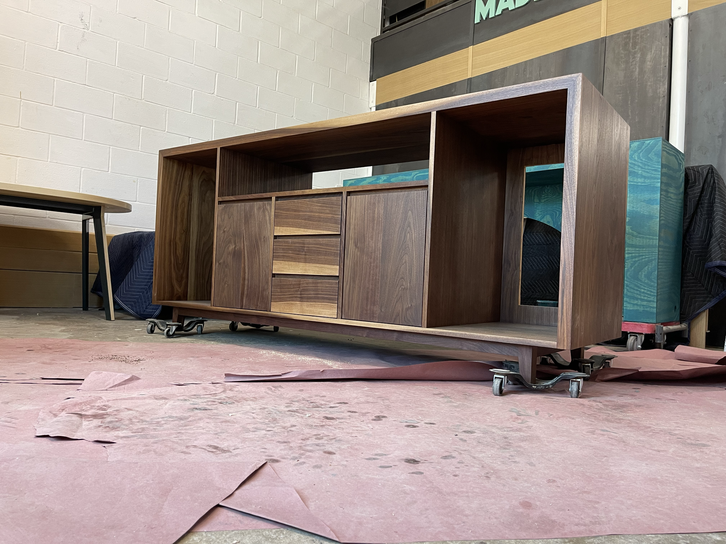 Wooden cabinet on caster wheels in a workshop setting, with pink paper or fabric on the floor and a white brick wall in the background.