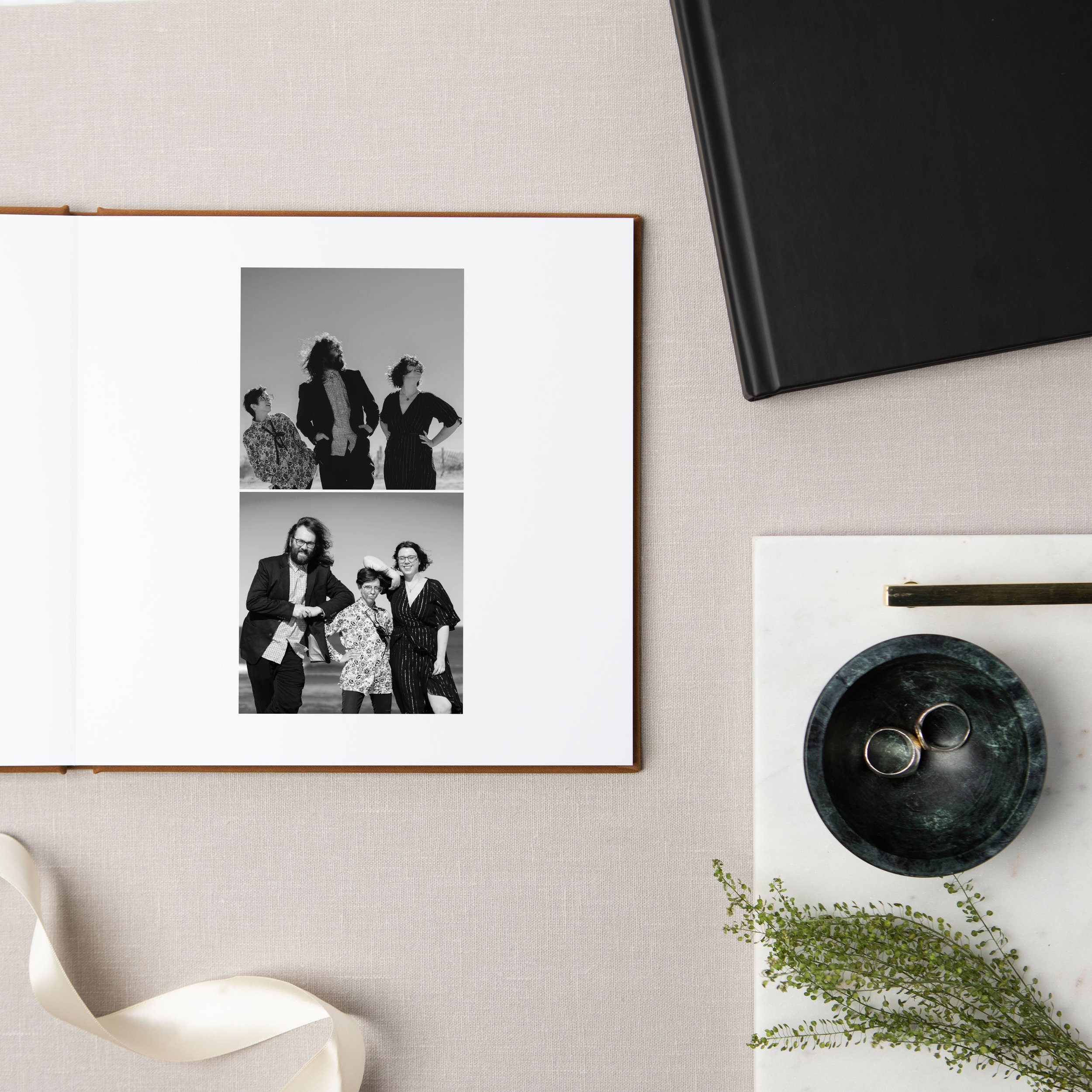 An open photo album displaying two black and white photos of a family at the beach, surrounded by a black notebook, a marble tray with rings, a small plant, and writing utensils on a neutral-colored desk.