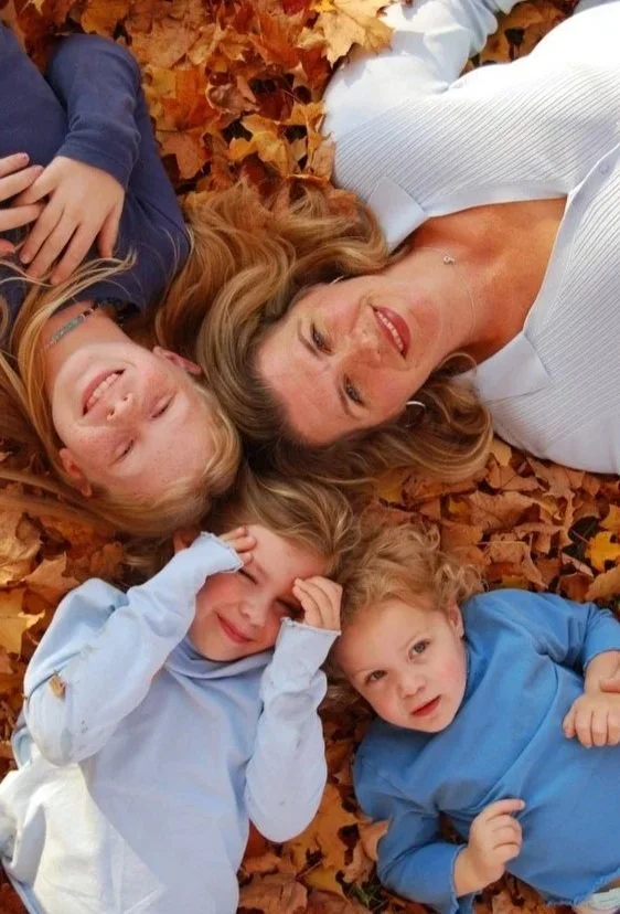 A woman and three children lying on a bed of autumn leaves, smiling and looking up at the camera.