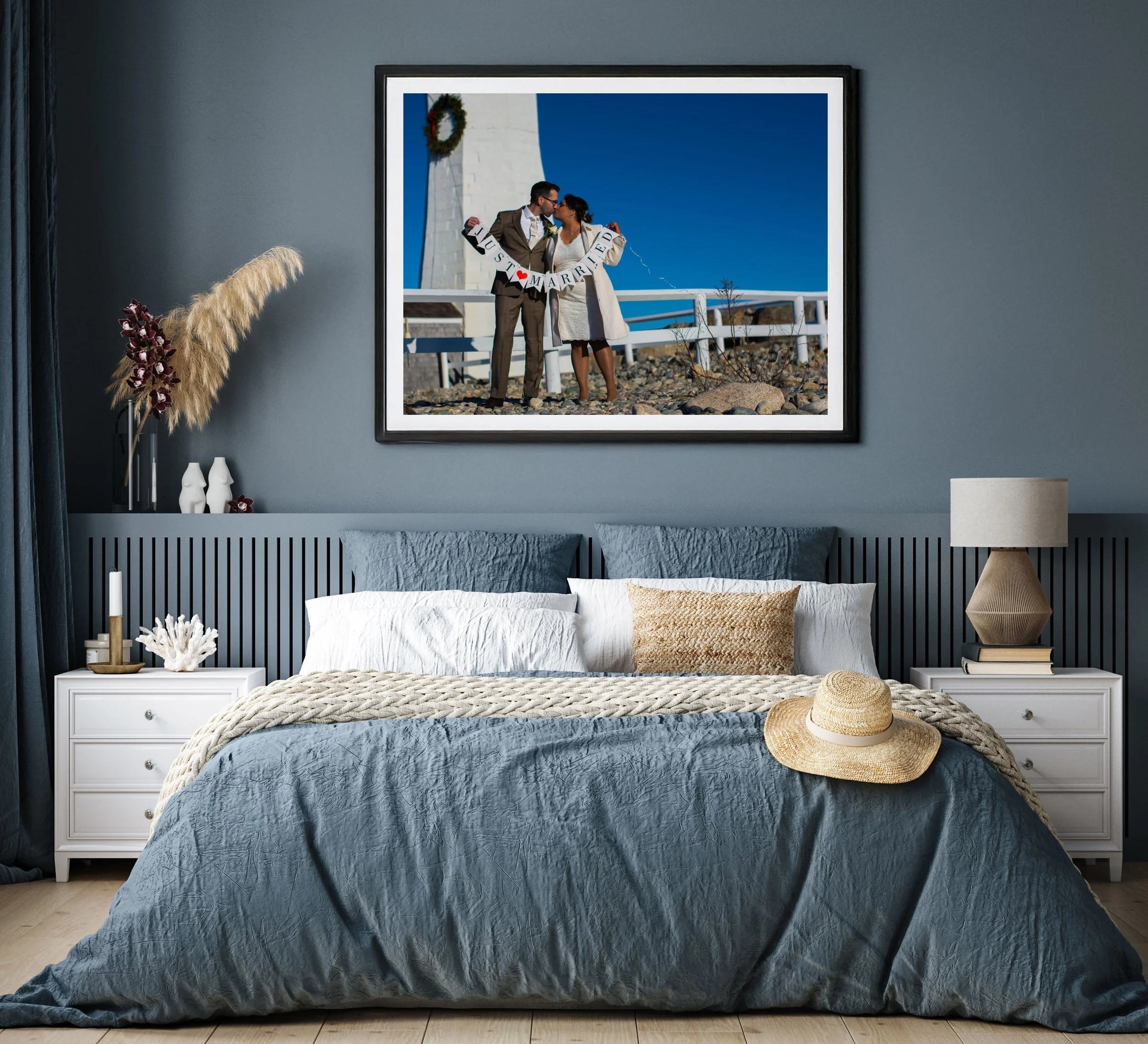 A decorated bedroom with a large framed wedding photo on the wall, two white nightstands with decorative items, a blue bed with white and beige pillows, a beige blanket, a straw hat on the bed, and a table lamp and books on the right nightstand.