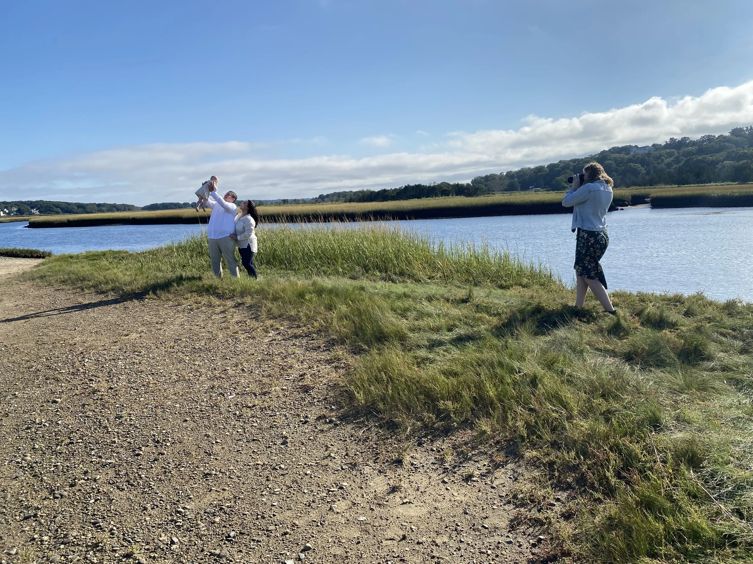 A family of three with a young child enjoying a day by the water, with a woman taking photos of them, on a sunny day with a clear blue sky and grassy landscape.