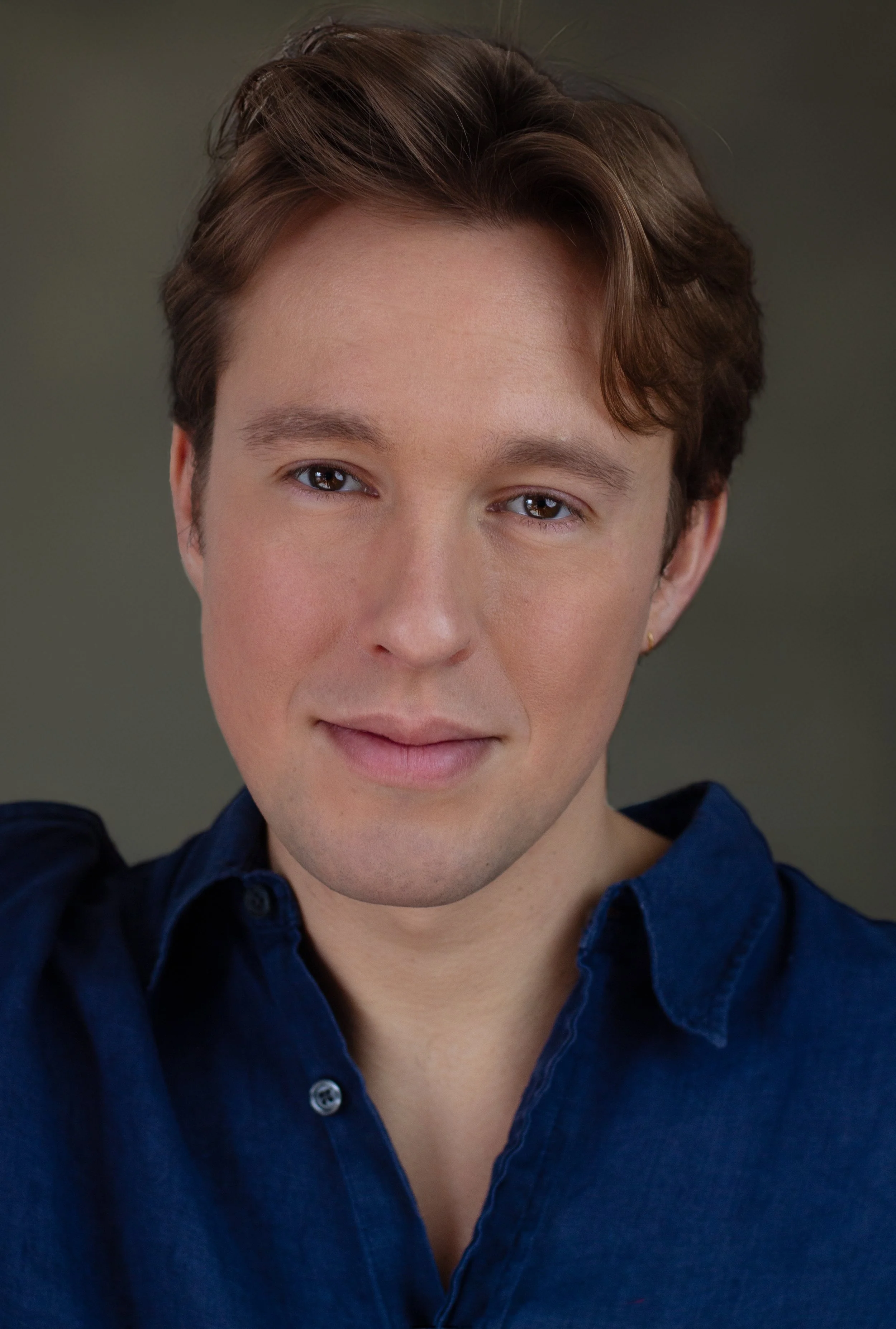 Close-up portrait of a person with short brown hair, wearing a dark blue collared shirt, smiling softly against a plain background.