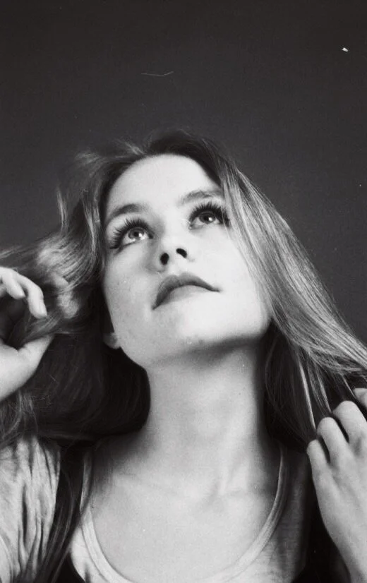 Black and white photo of a young woman looking upward, holding her hair, with long wavy hair and a contemplative expression.