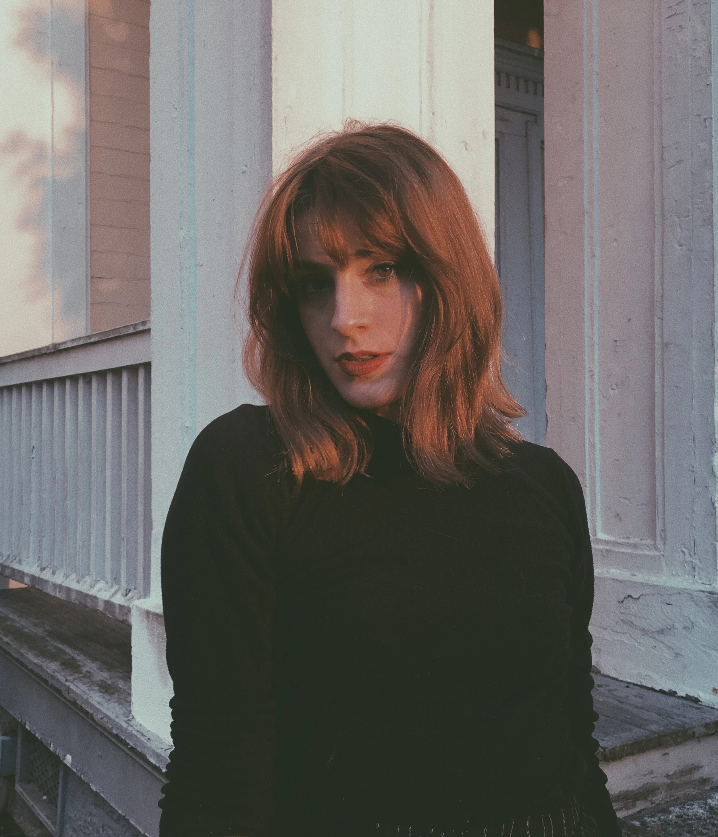 A woman with shoulder-length reddish-brown hair wearing a black top, standing outside a white house with wooden siding and a railing, during sunset.