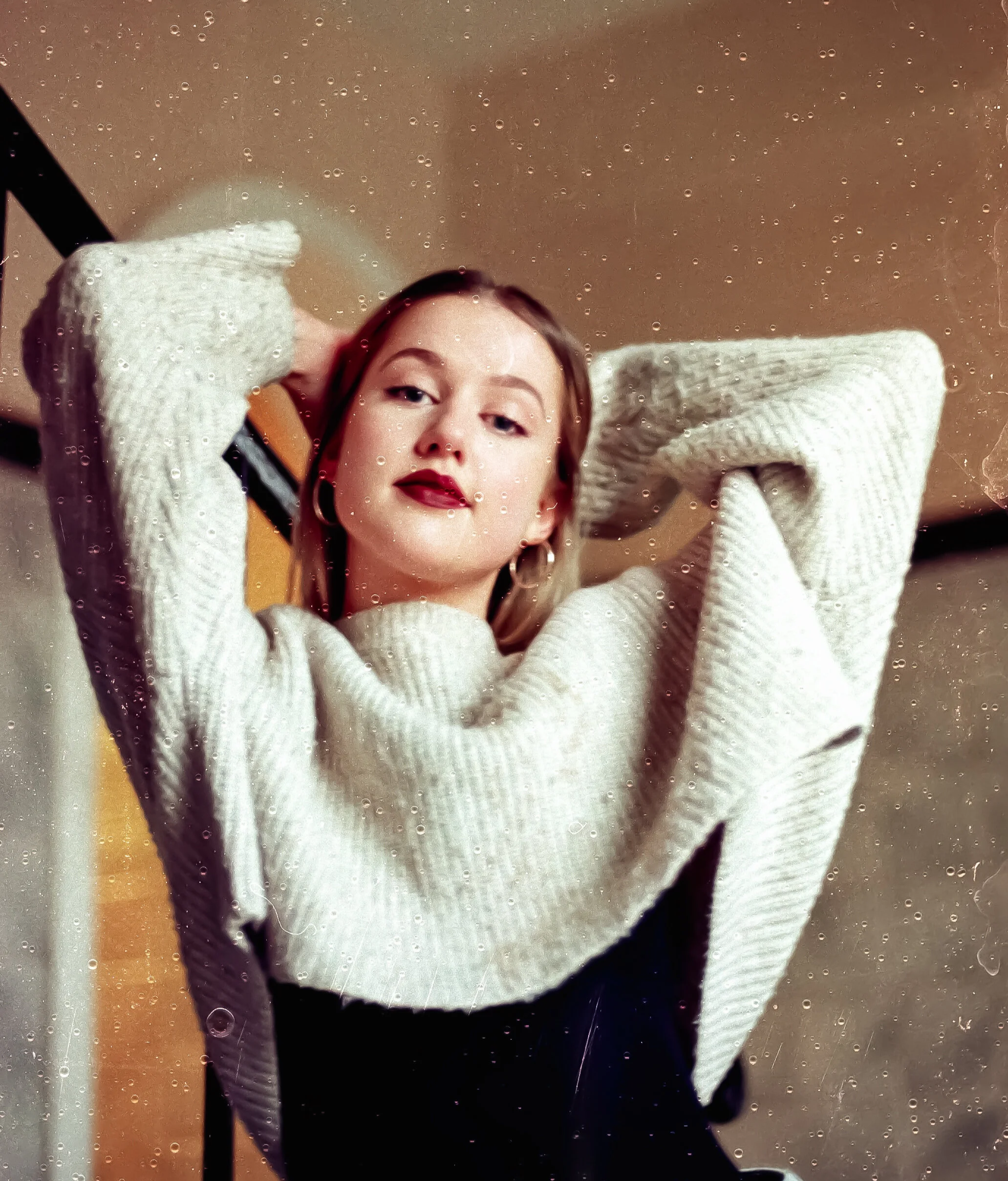 Portrait of a young woman with fair skin, wearing a white sweater and black pants, posing with her hands behind her head, looking at the camera with a confident expression, through a window with water droplets.