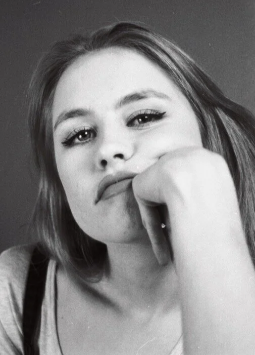 Close-up of a young woman with shoulder-length hair, resting her chin on her hand, looking at the camera with a neutral expression.