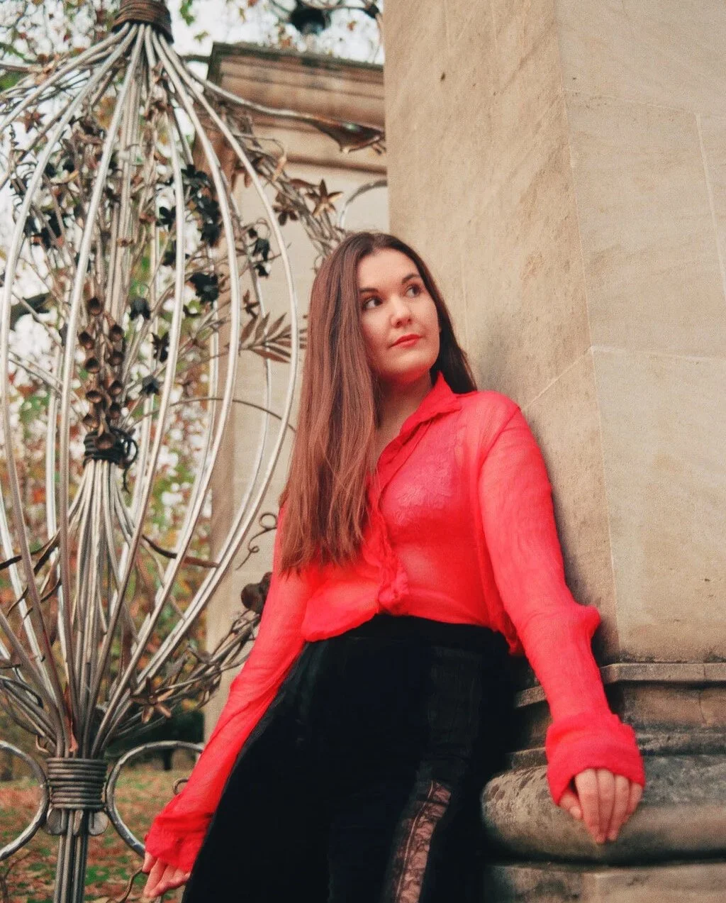 Young woman with long brown hair leaning against a stone column outdoors, wearing a red sheer blouse and black pants, next to metal decorative sculpture and plants.