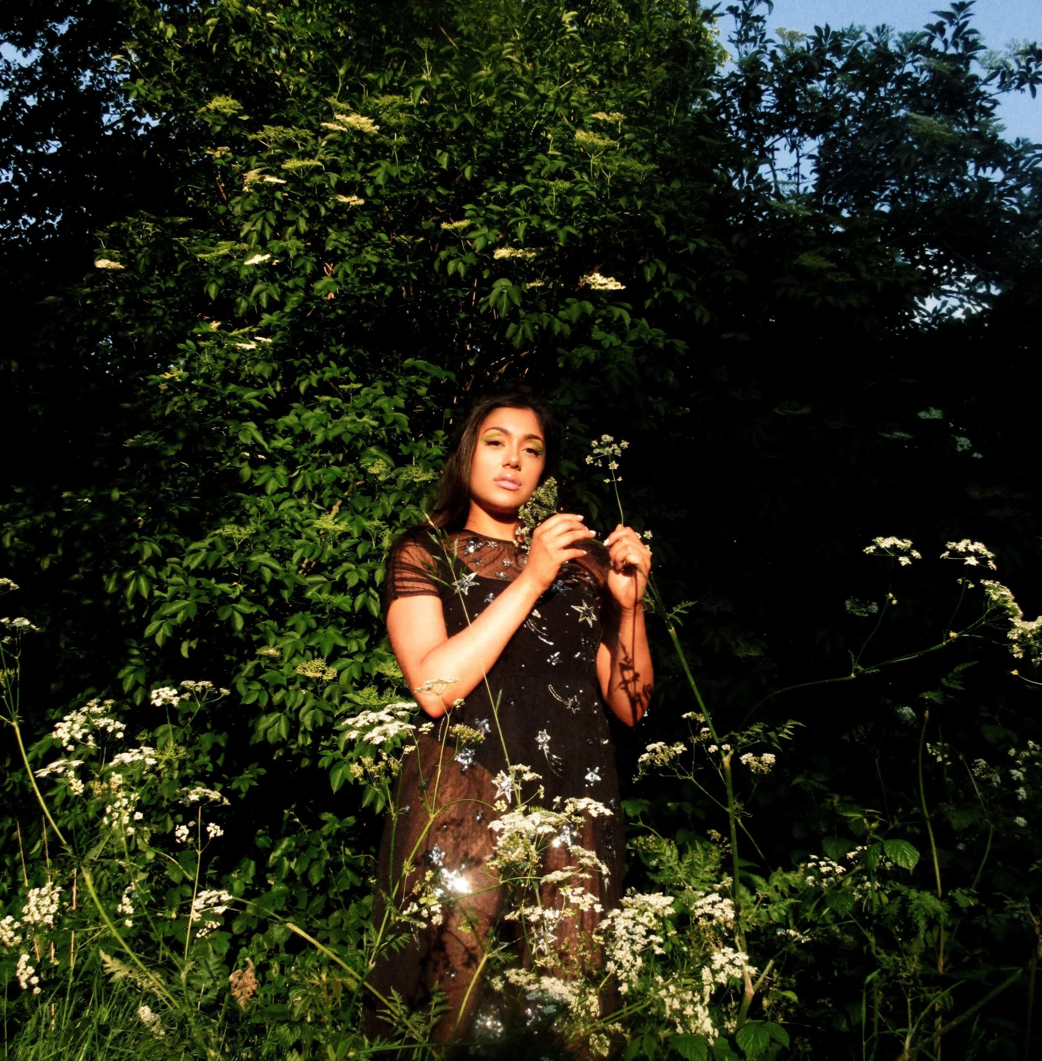 A woman with long dark hair is standing outdoors at dusk, surrounded by green foliage and white wildflowers. She is wearing a sheer black dress with star and celestial patterns, and is holding a small flower in each hand, looking at the camera with a contemplative expression.
