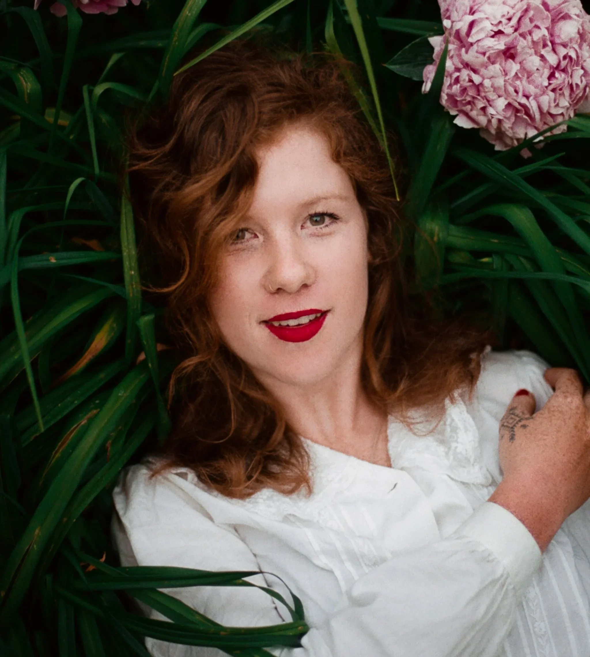 Woman with red curly hair lying on green grass next to pink flowers, wearing a white blouse and red lipstick.