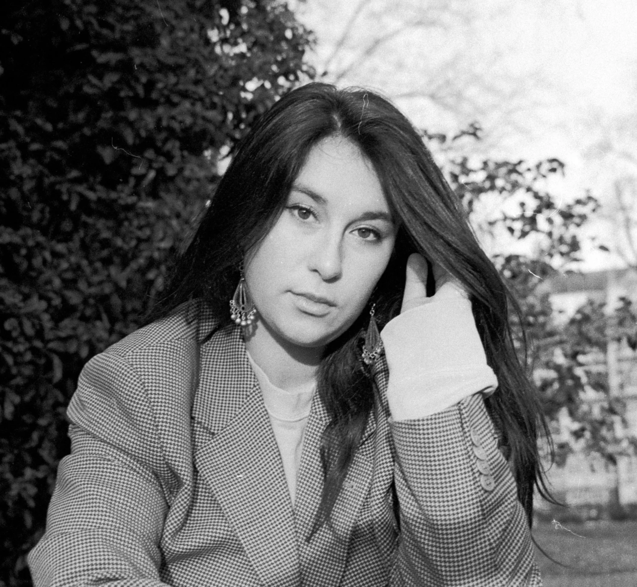 Black and white photo of a young woman with long dark hair, wearing earrings and a checkered blazer, outdoors with trees and houses in the background.