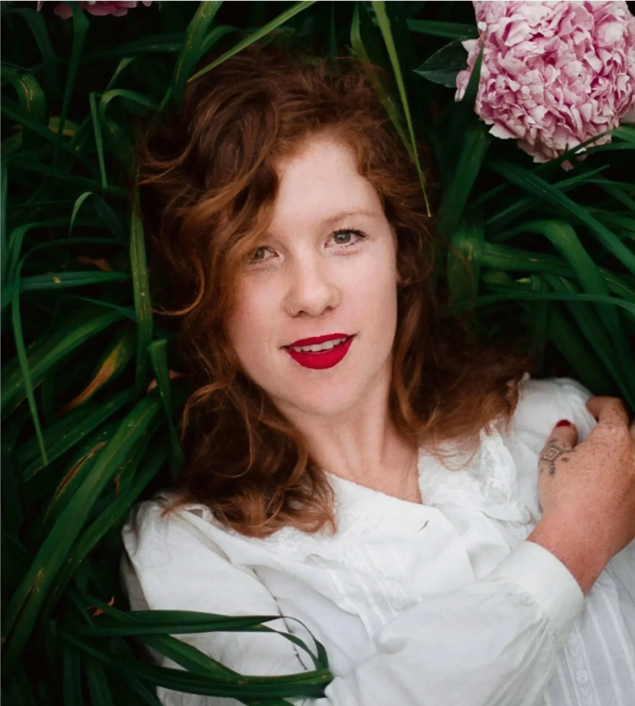 A woman with red curly hair lying among green plants and pink flowers, wearing a white blouse with red lipstick, smiling at the camera.