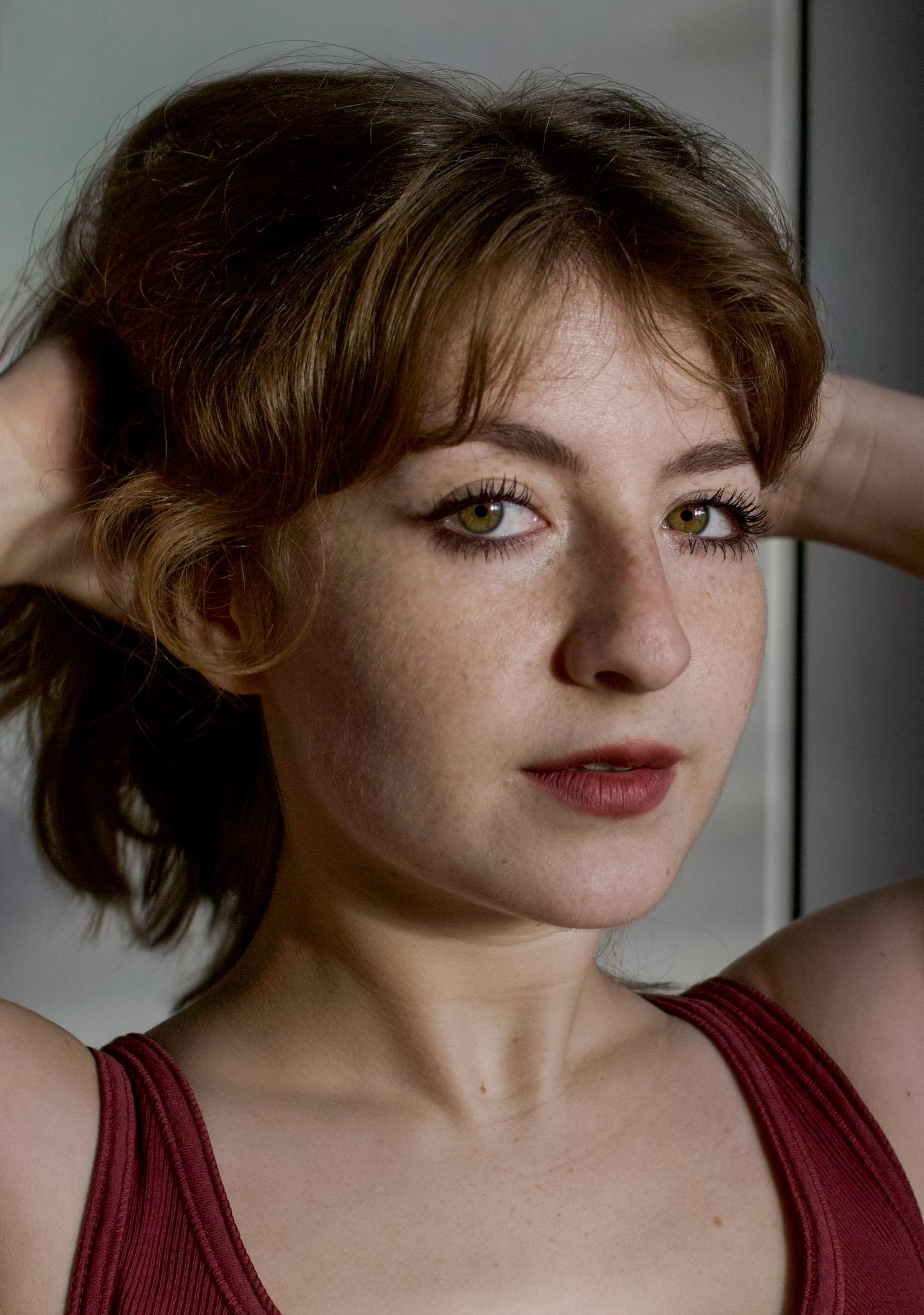 A young woman with curly red hair and green eyes, looking at the camera with her arms raised behind her head, wearing a red tank top.
