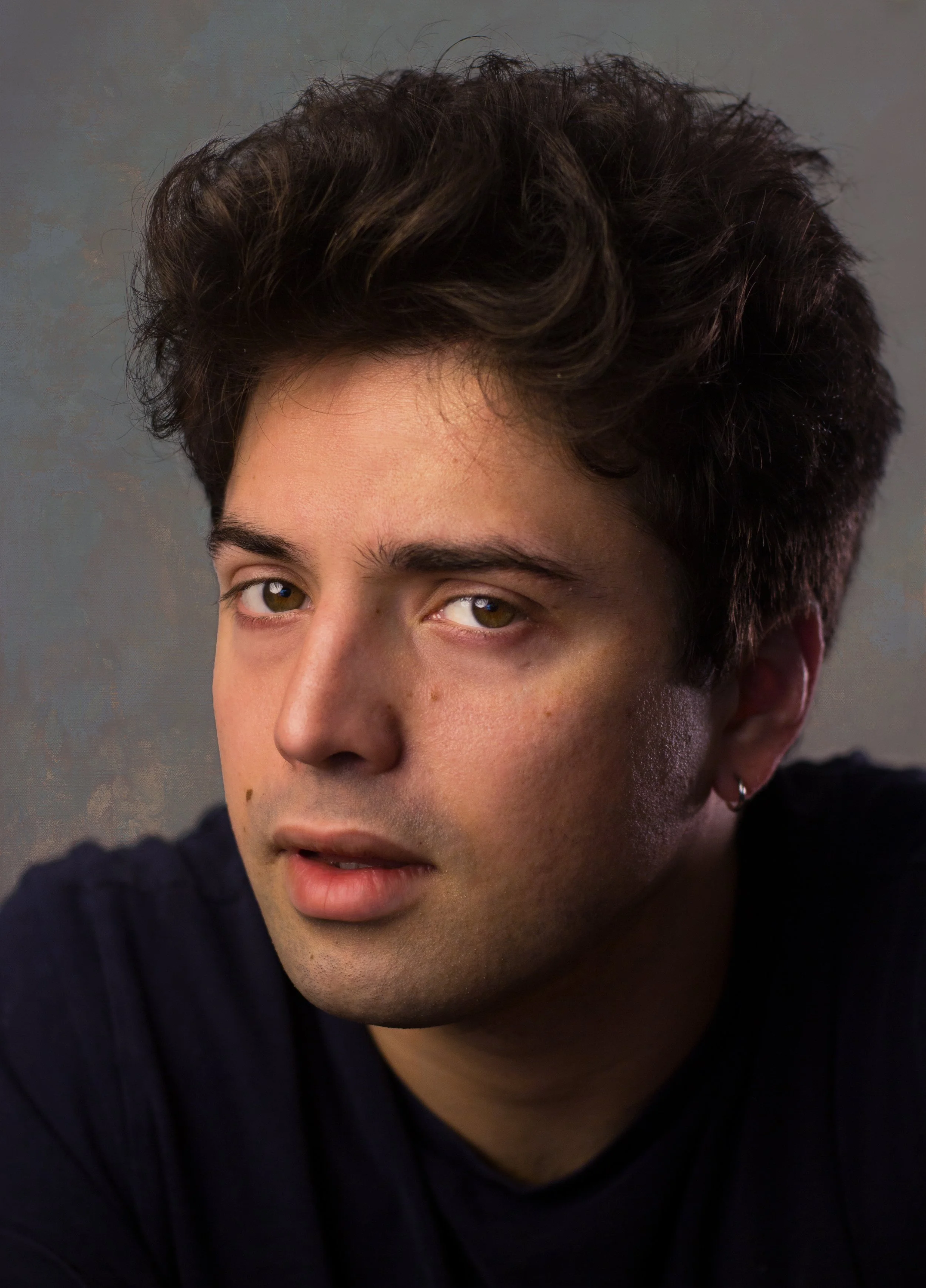 Close-up portrait of a young man with curly dark brown hair, green eyes, and an earring, wearing a black shirt, against a neutral background.