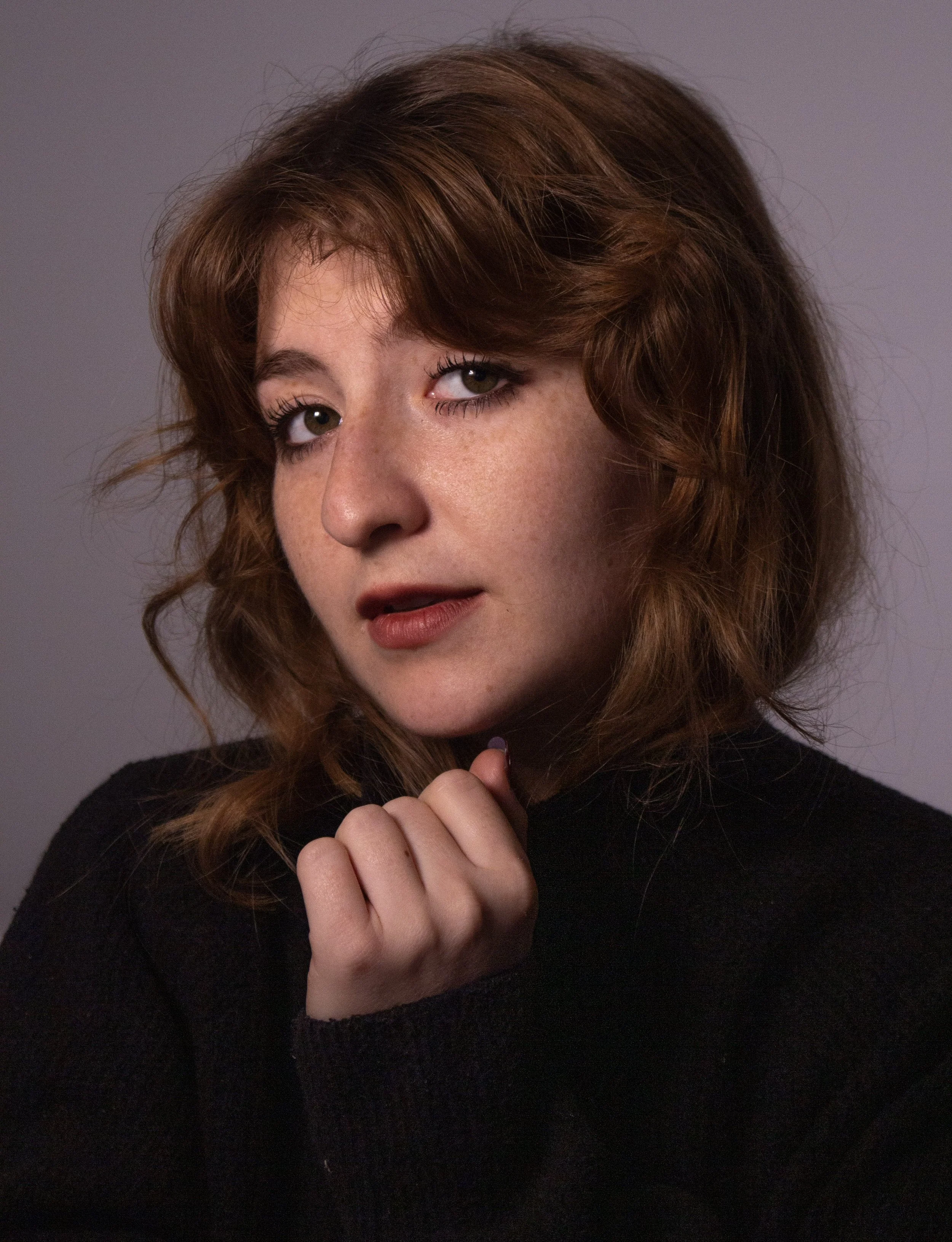 A woman with wavy red hair and green eyes, wearing a black top, looking at the camera with a relaxed pose against a plain gray background.