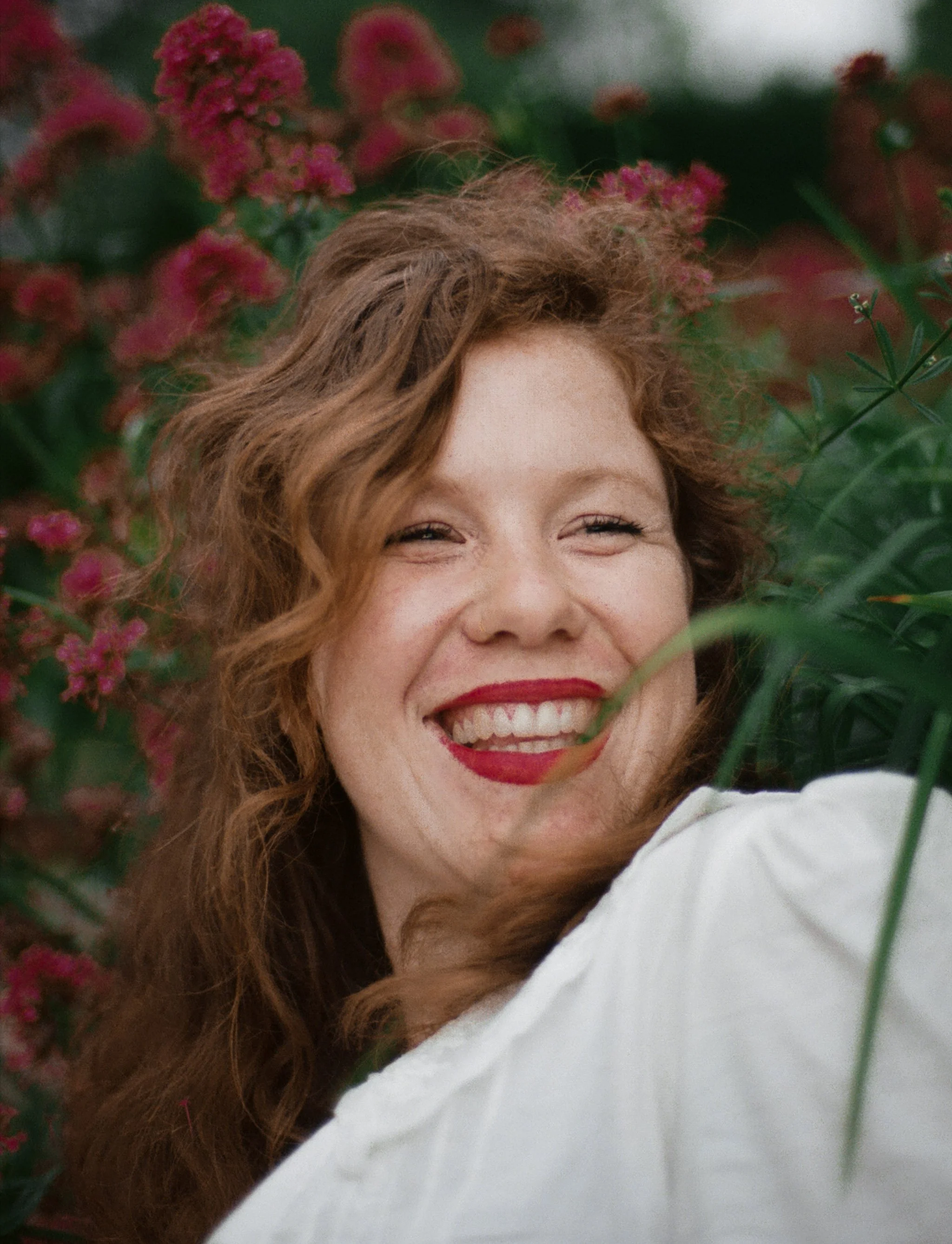 A woman with curly red hair and bright red lipstick smiling among pink flowers and green foliage.