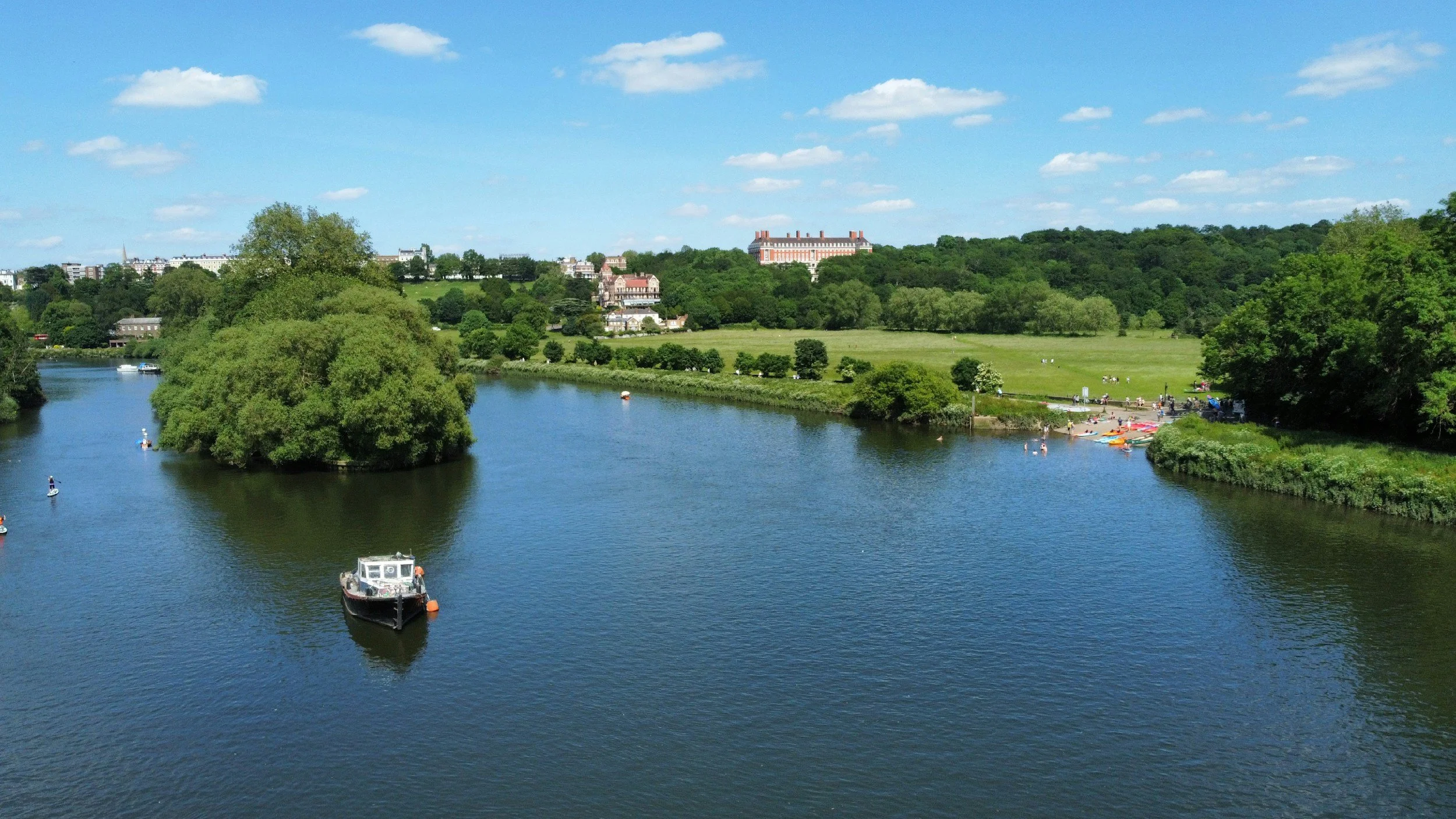 A river flowing through a green landscape with trees, grass, and people relaxing on the riverbank, along with boats on the water, under a blue sky with scattered clouds.