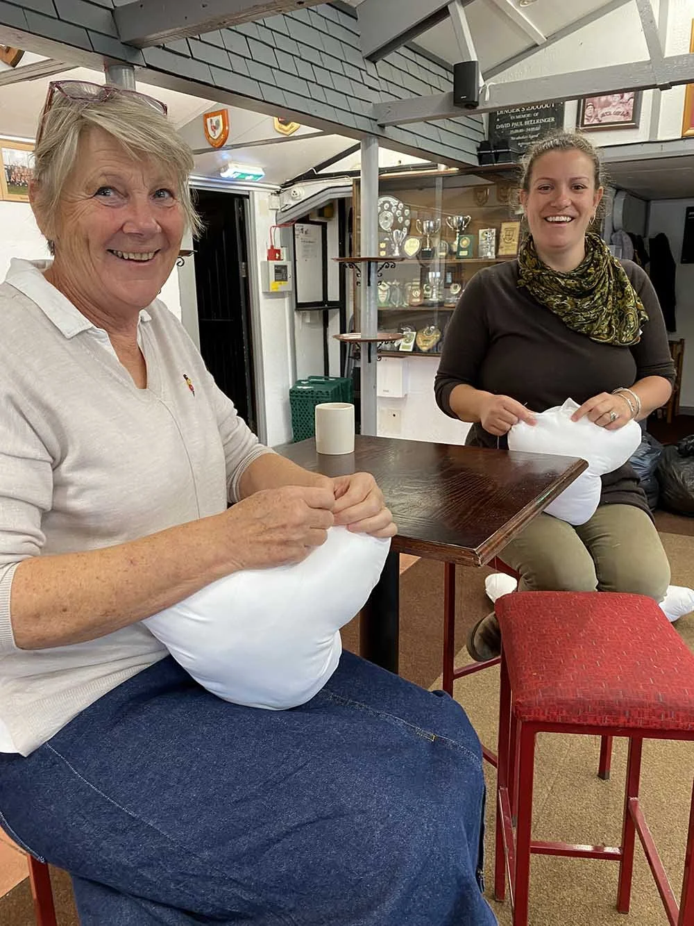 perfect Hug sewing team: Two women sitting at a table, holding white heart pillows and smiling.