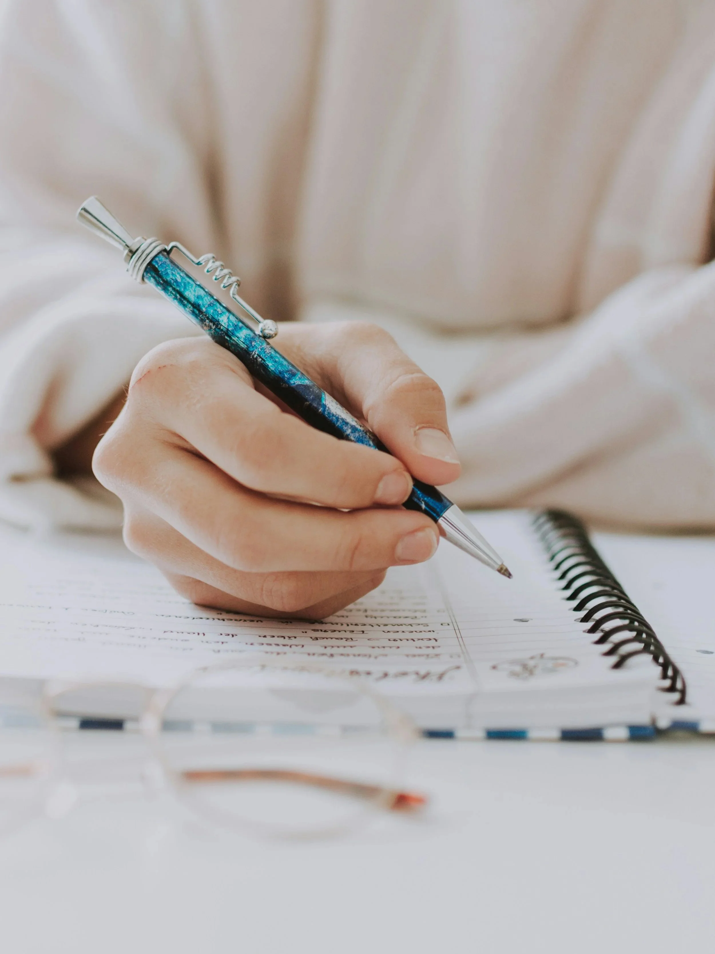 Close-up of a person writing in a spiral notebook with a blue and silver pen, with blurred white background.