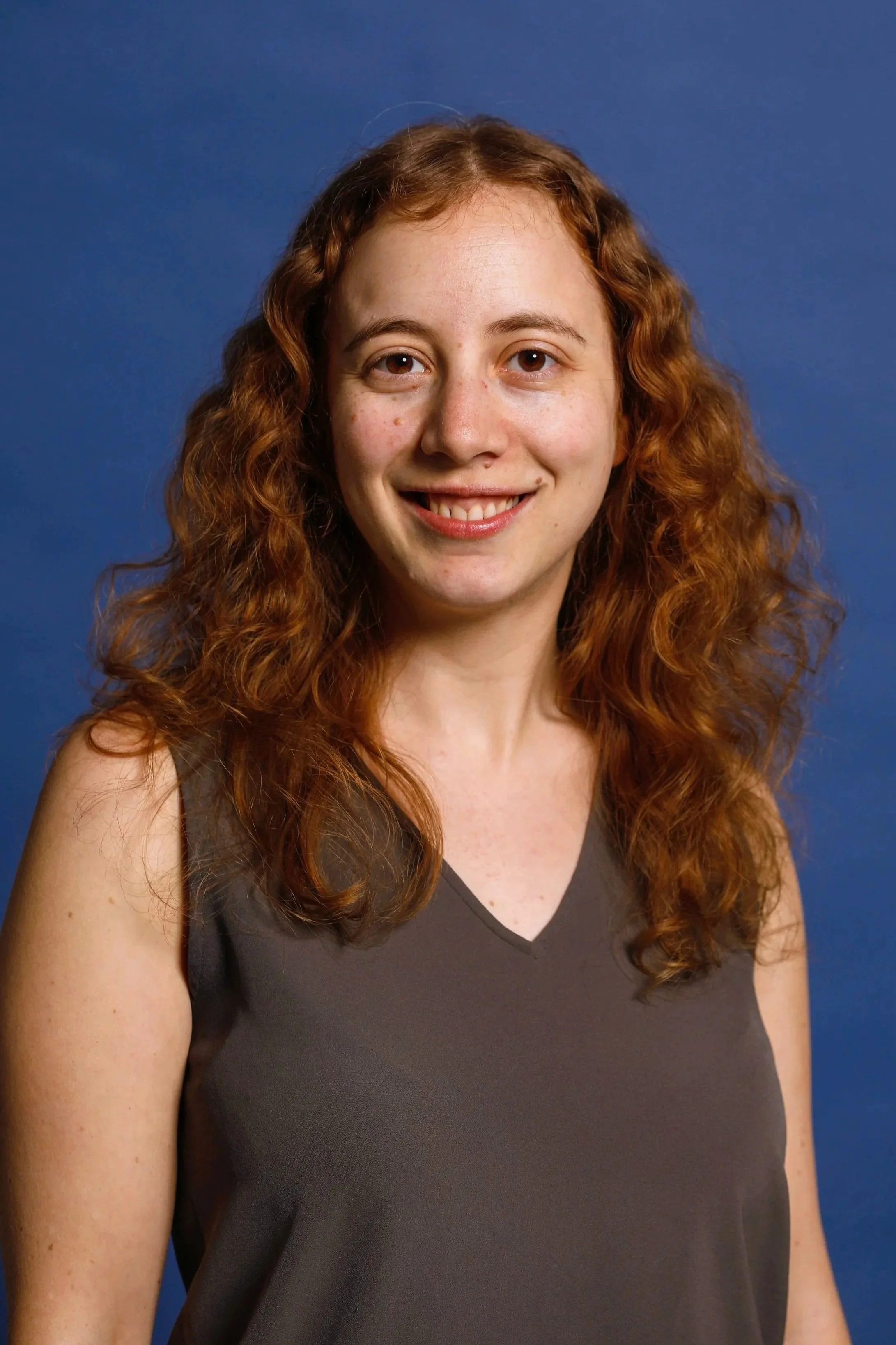 A woman with curly red hair smiling, wearing a sleeveless dark gray top, against a plain blue background.